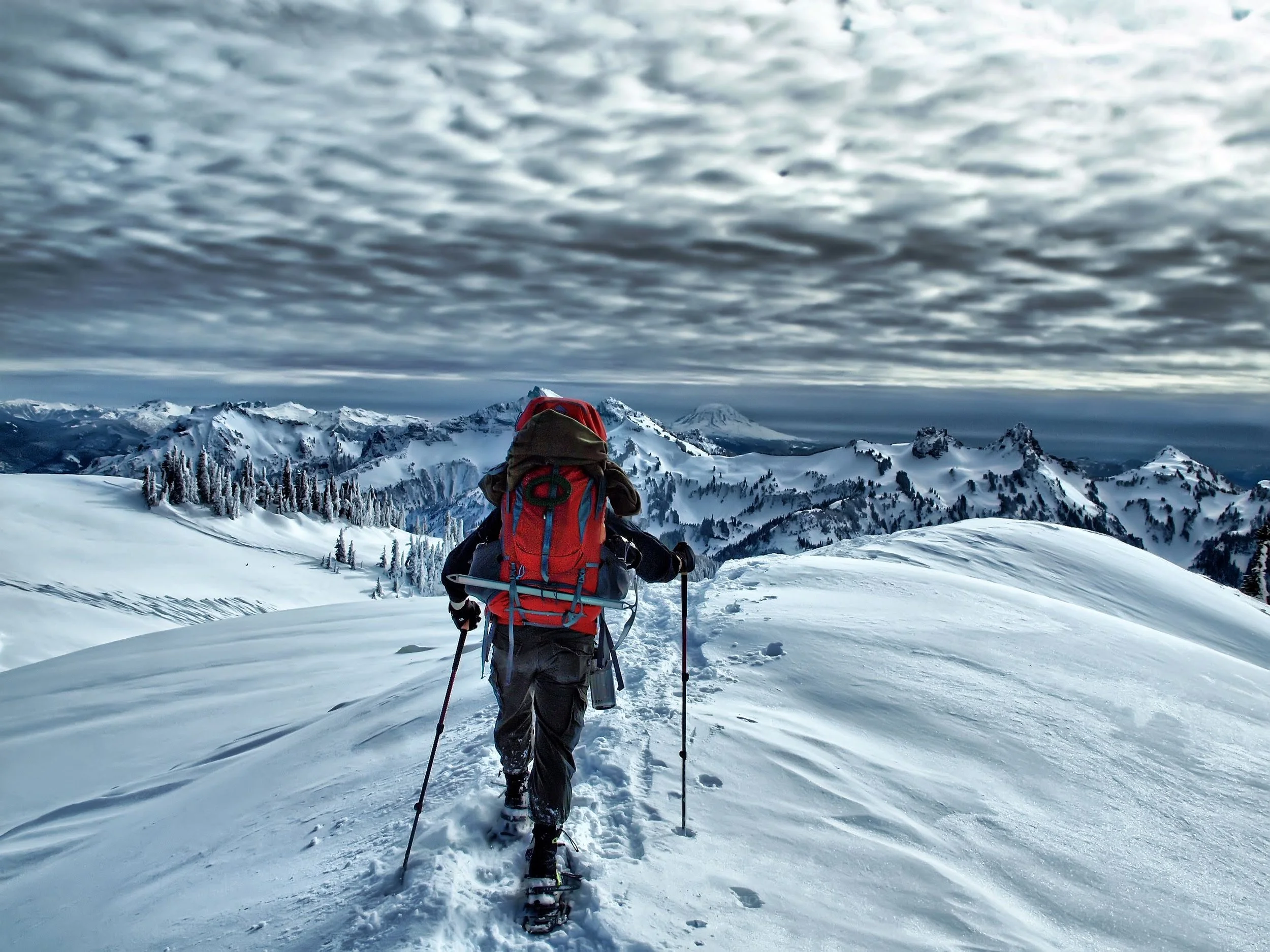 A person hiking in a snowy mountain landscape wearing a red jacket and carrying a large backpack, with snow-covered mountains and a cloudy sky in the background.