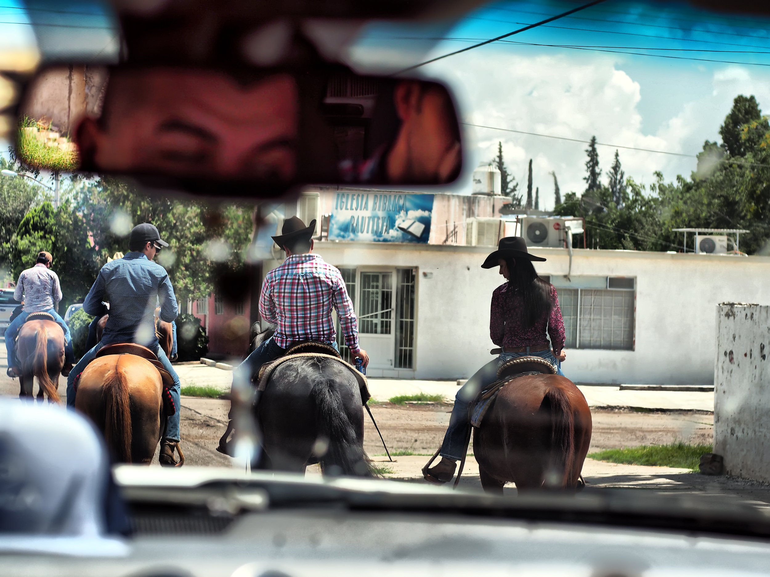 View of three people riding horses down a street, seen from inside a vehicle with the rearview mirror partially reflecting the person's eyes and forehead.