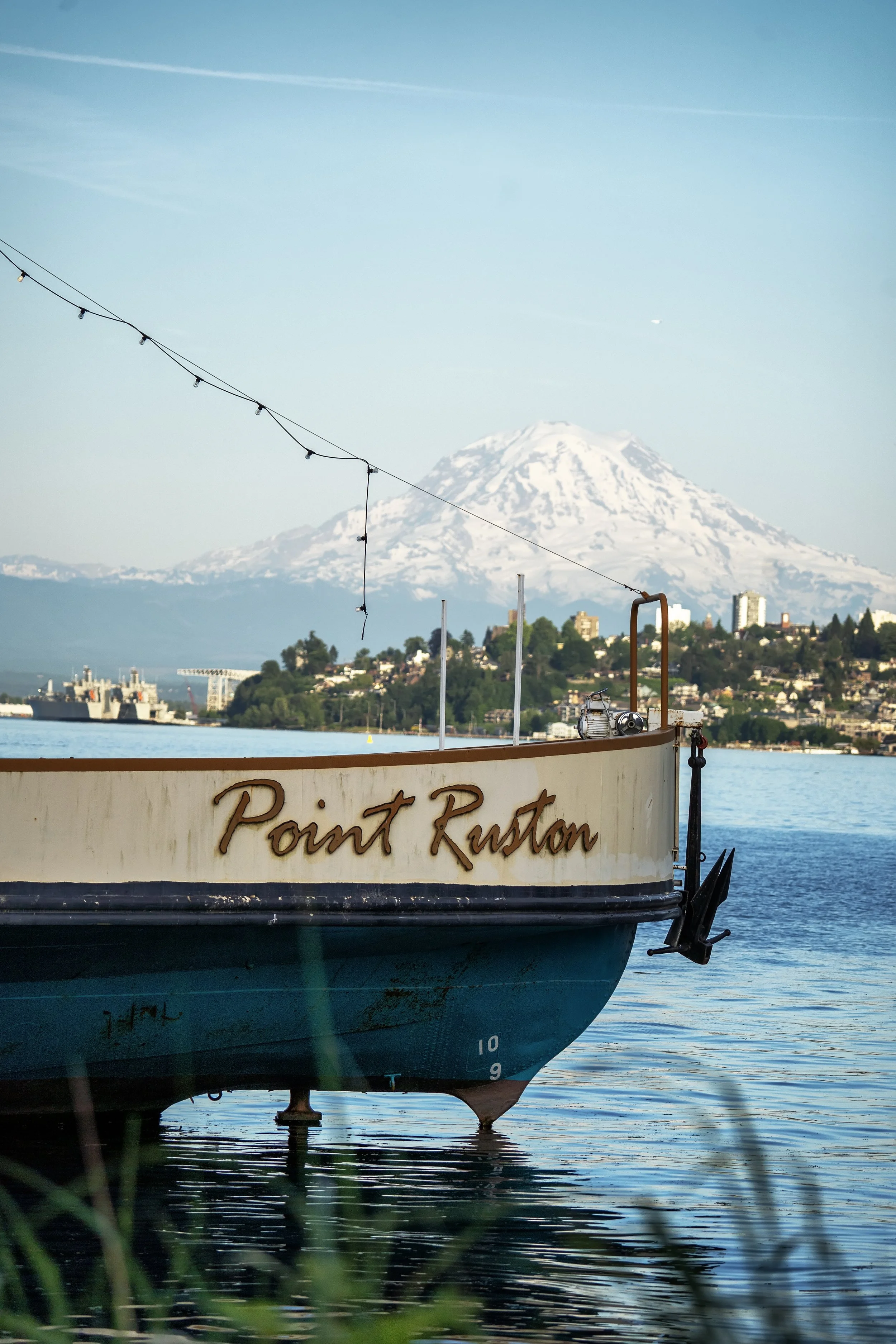 A boat named 'Point Ruston' moored in water with Mount Rainier in the background, buildings on a hillside, and string lights overhead near the boat.