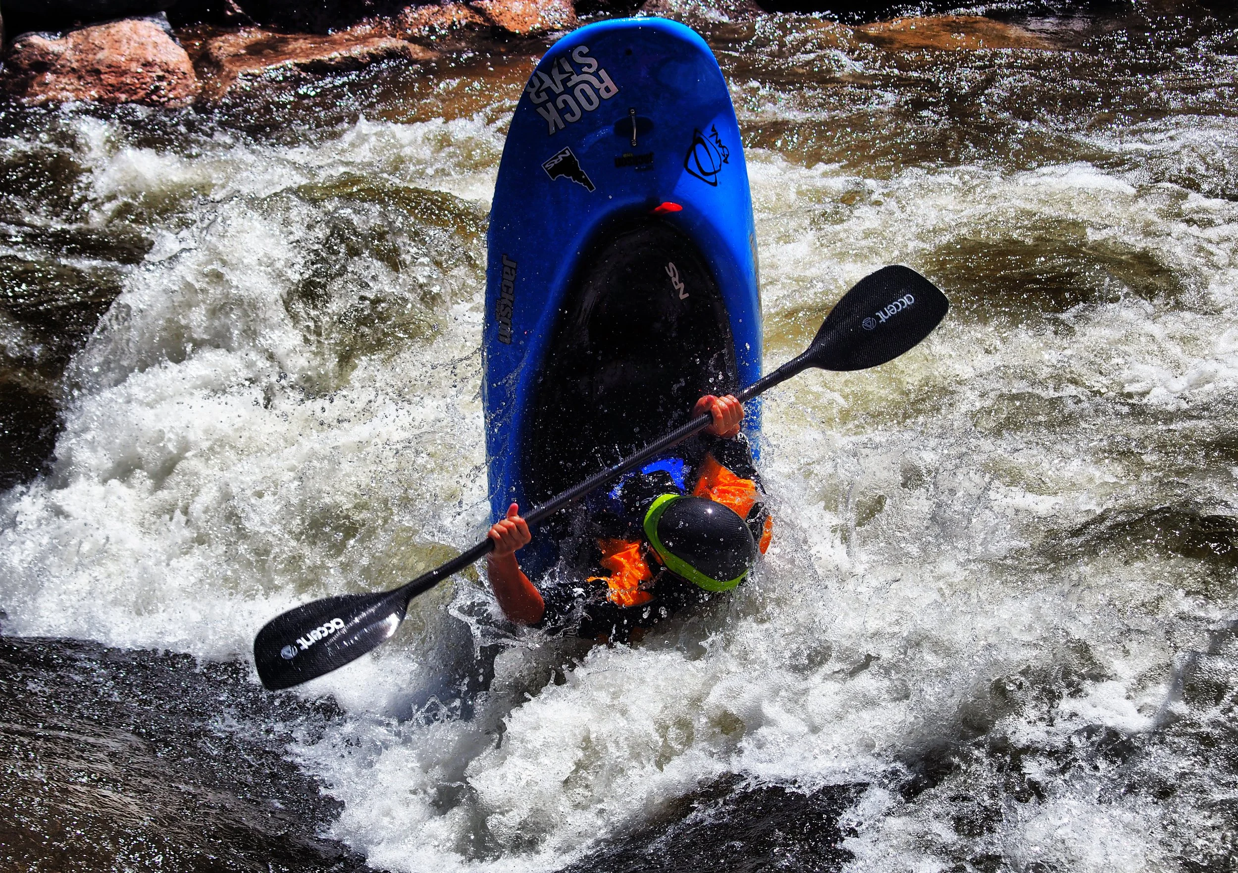 Person kayaking in rough water with foam, wearing a helmet and orange lifejacket, holding a paddle.