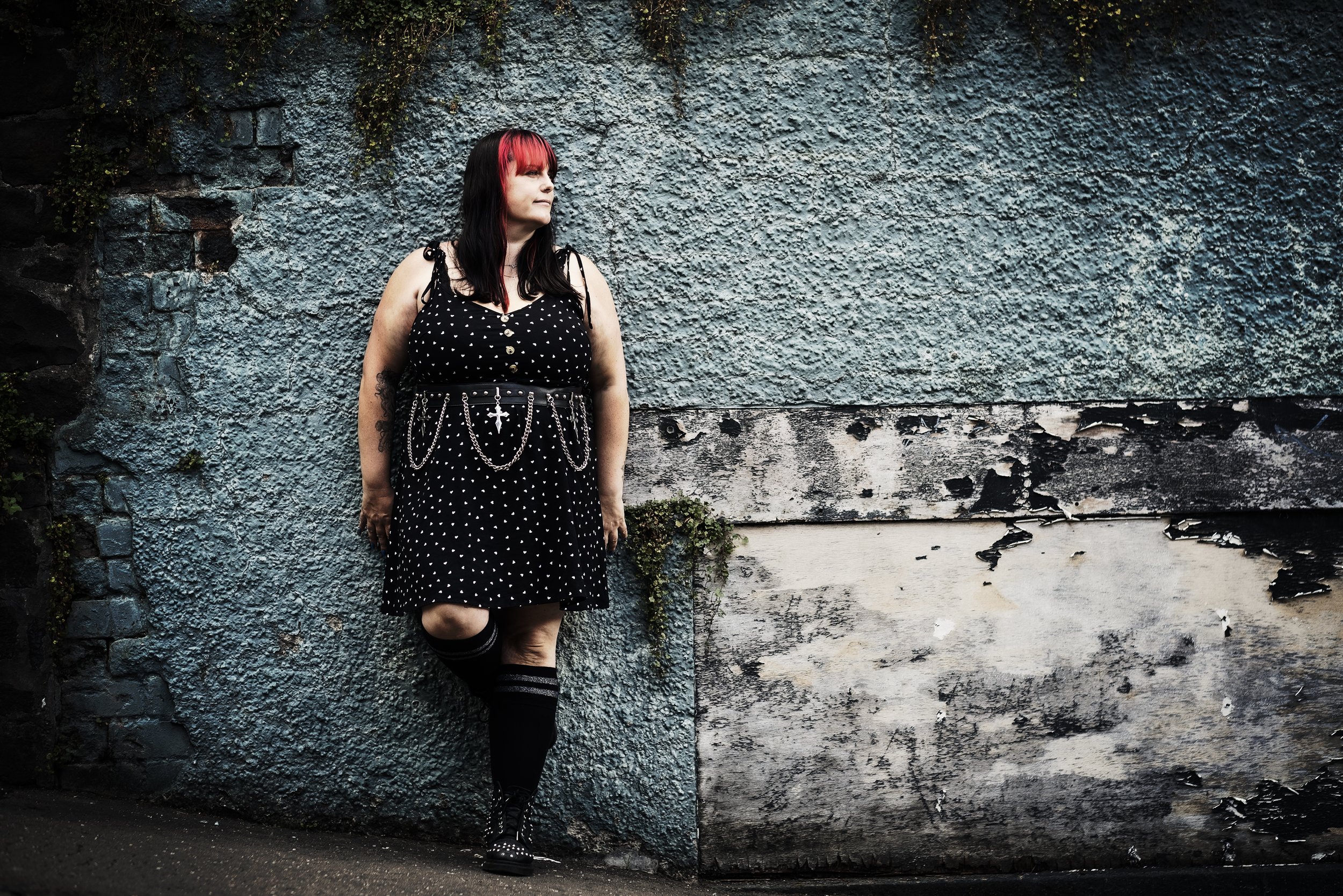 A woman with black and red hair, wearing a black polka dot dress with chains and a cross accessory, leaning against a textured gray and black wall with peeling paint.