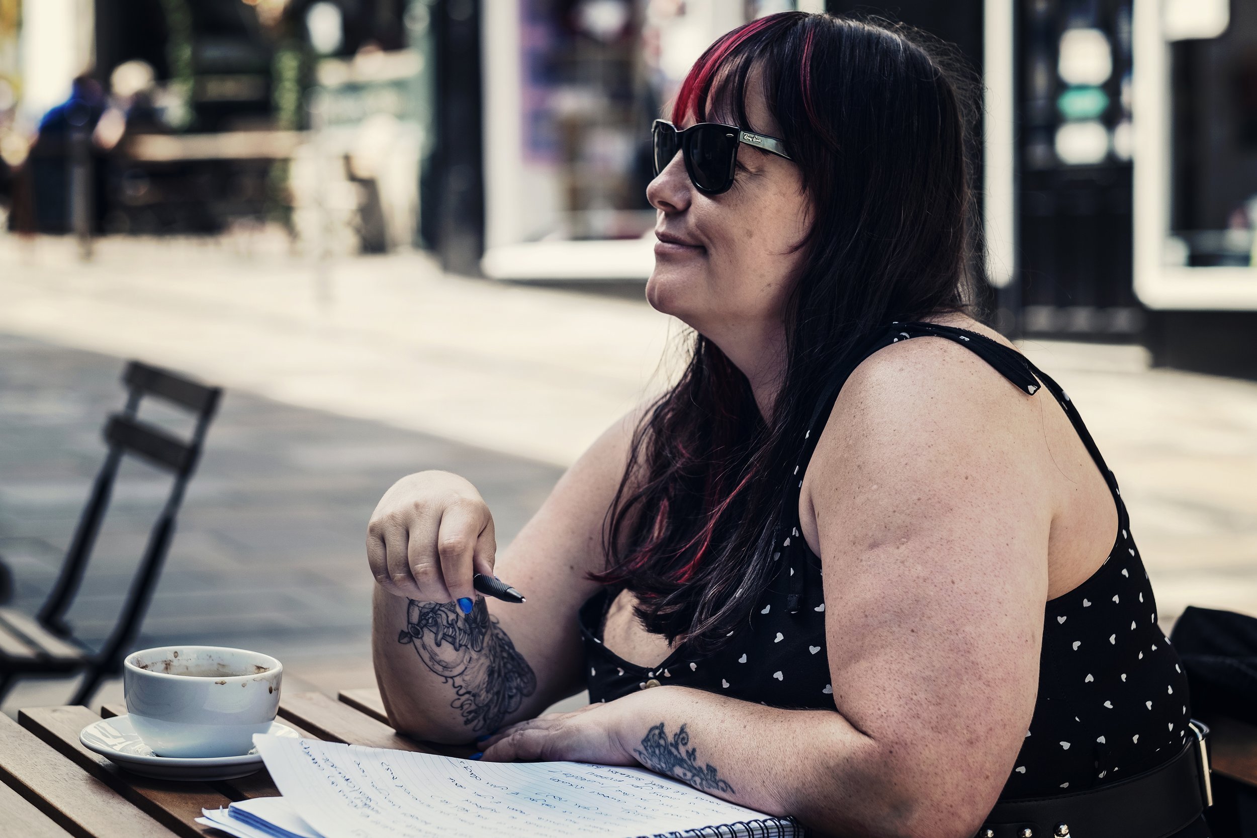 A woman with long dark hair, red highlights, and sunglasses sitting at an outdoor table with a cup of coffee, holding a pen over a notebook, wearing a black sleeveless dress with white heart patterns.