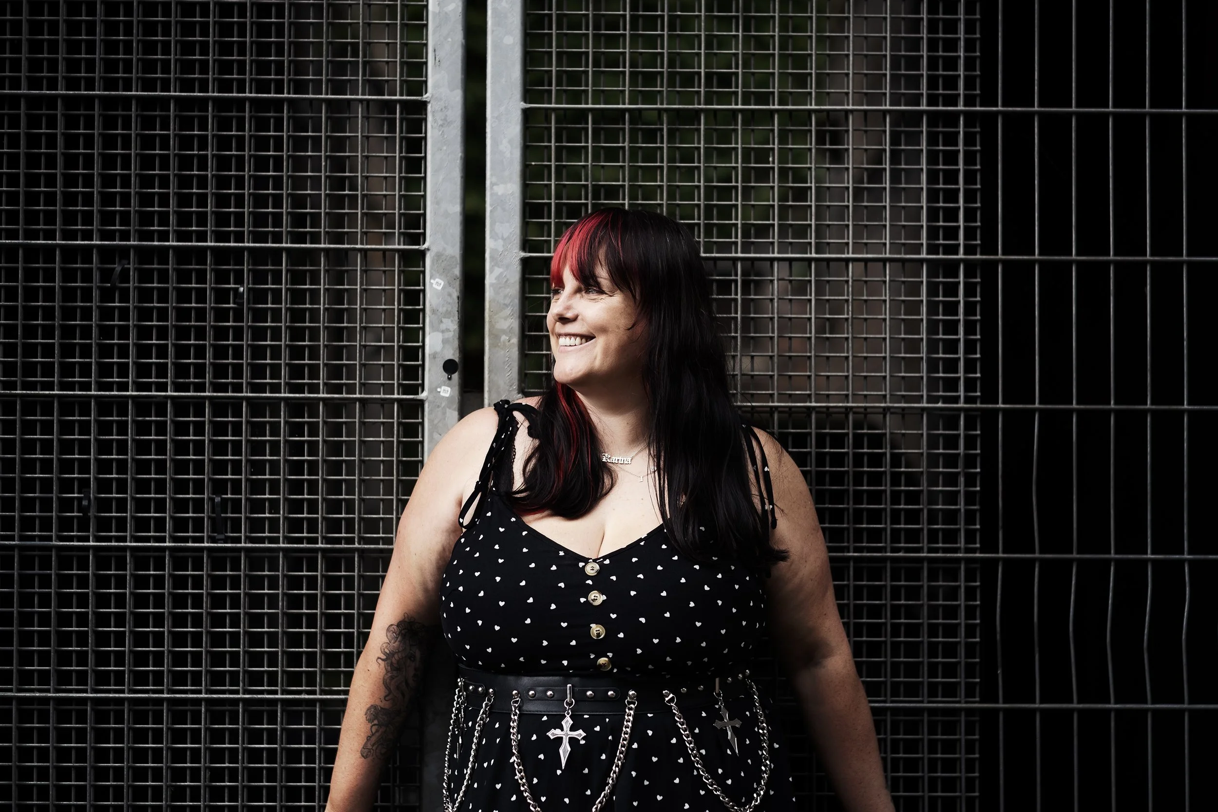 A woman with black and red hair smiling and looking to her left, standing in front of a metal fence.
