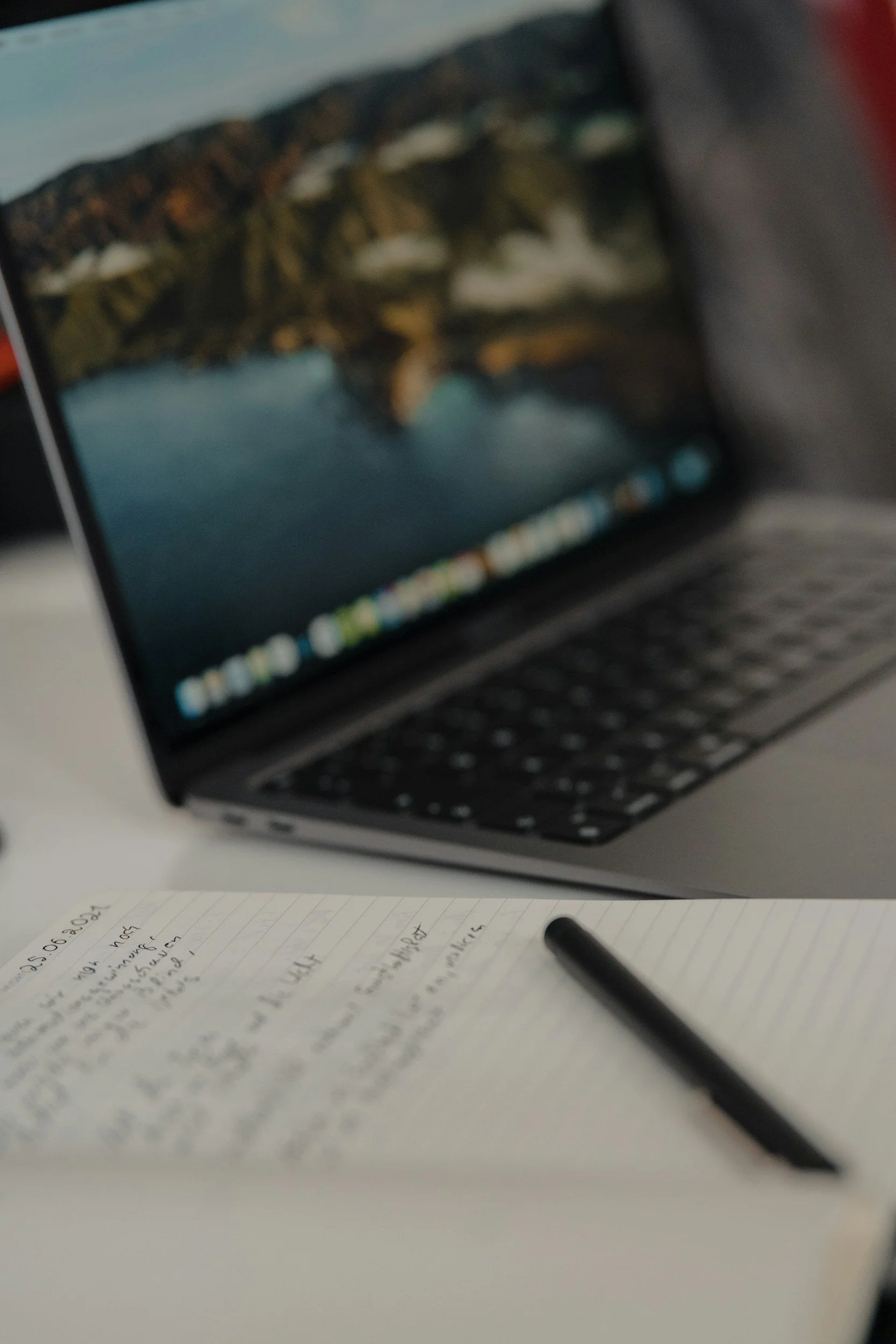 A notebook with handwritten notes and a pen resting on it, placed on a white surface, with a laptop in the background displaying a blurry scenic landscape.