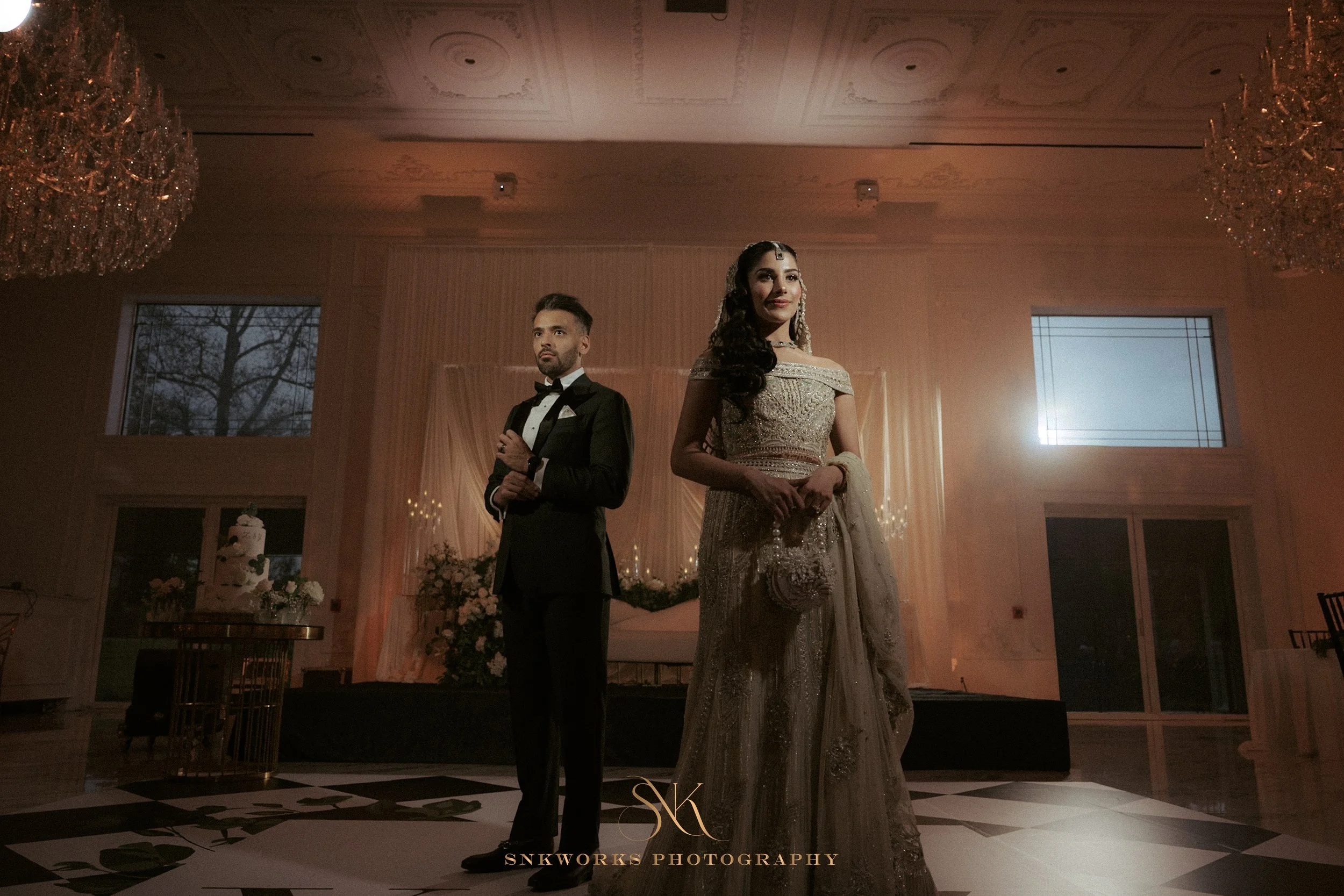 A bride and groom standing together at their wedding reception, dressed in traditional and formal attire, with elegant decor and a chandelier in the background.