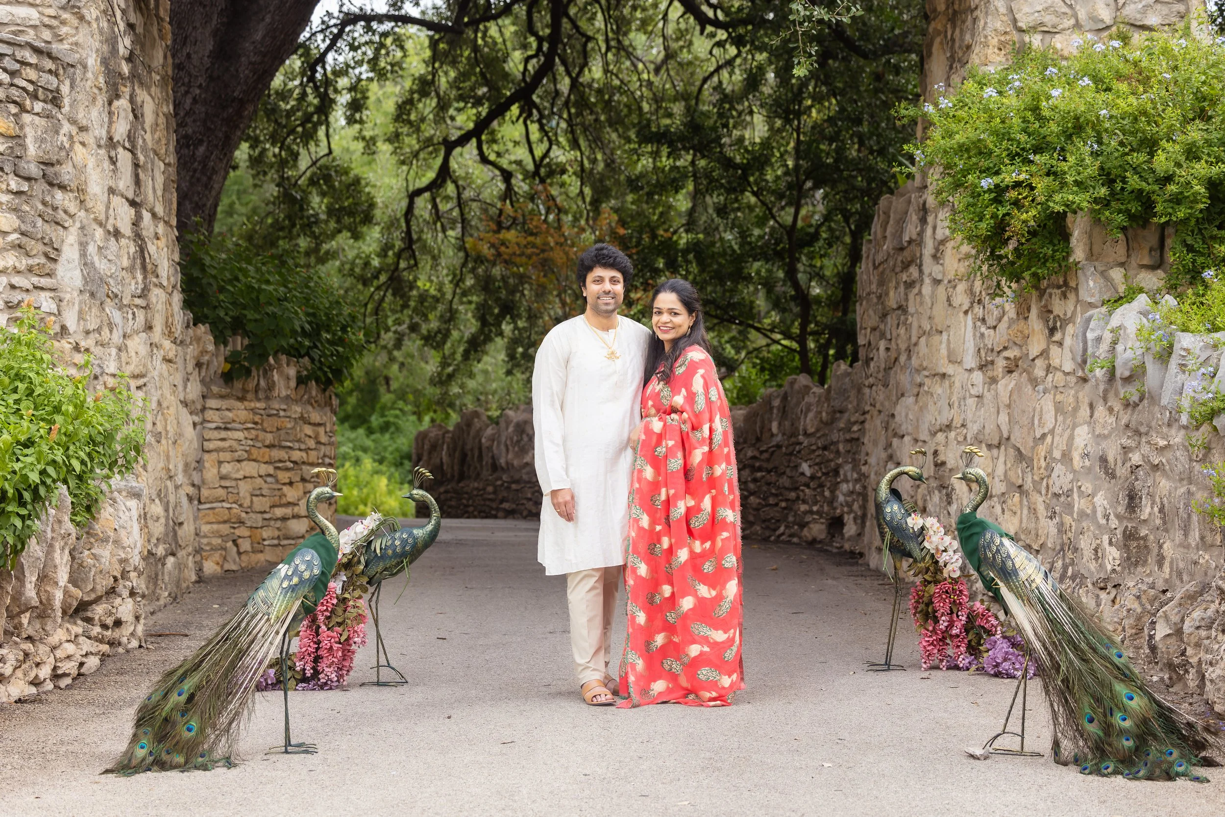 A smiling couple standing on a pathway decorated with peacock sculptures and flowers, surrounded by stone walls and lush green trees.