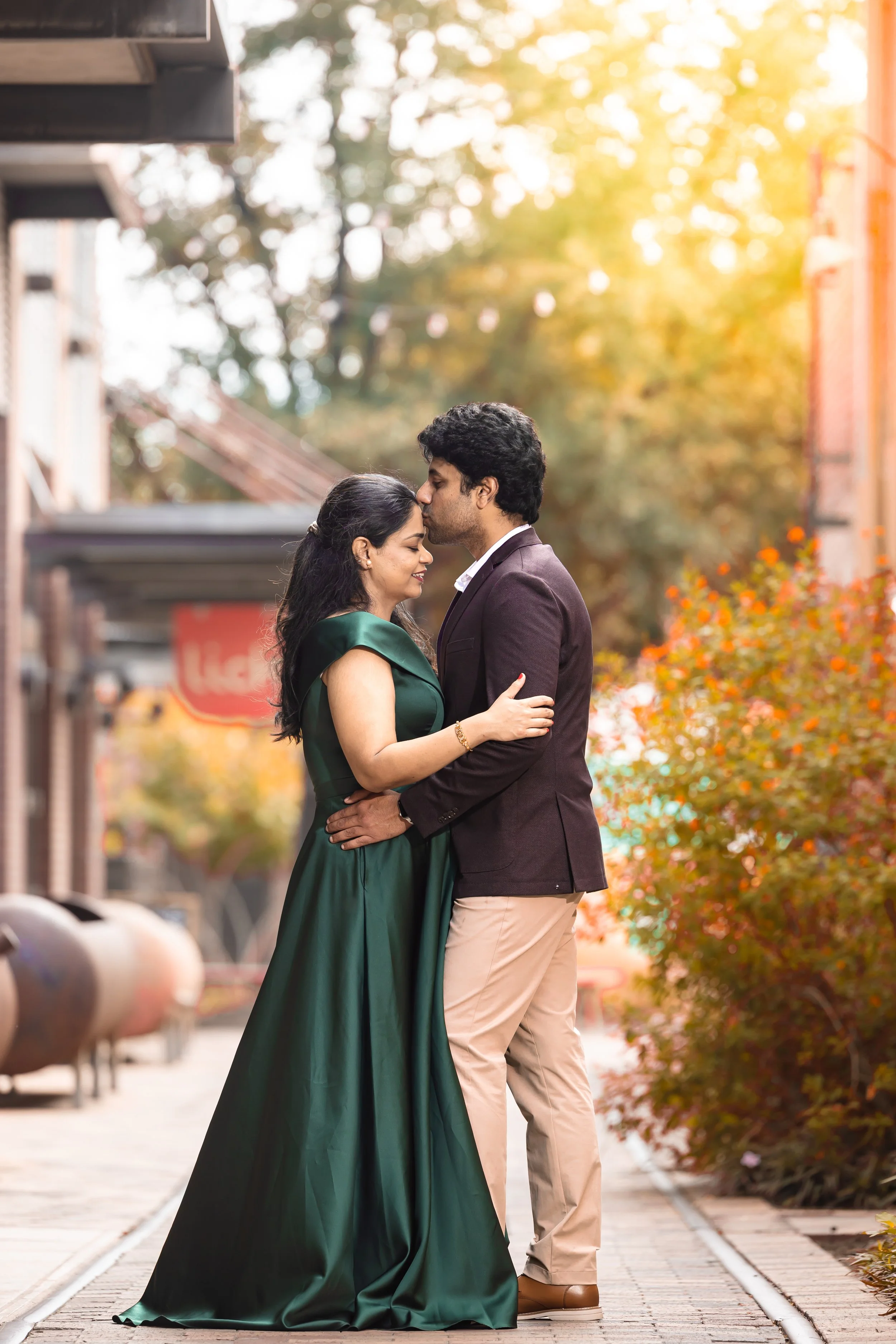 A romantic couple embracing on a city street during golden hour, with trees and buildings in the background.