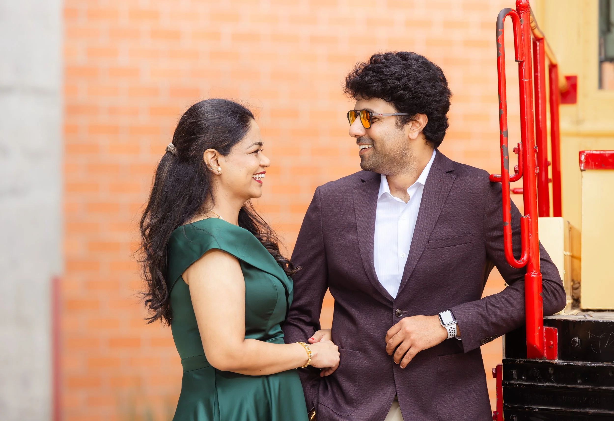 A smiling man and woman face each other, holding hands, outdoors near a brick wall and yellow and red metal structure, dressed in formal attire.