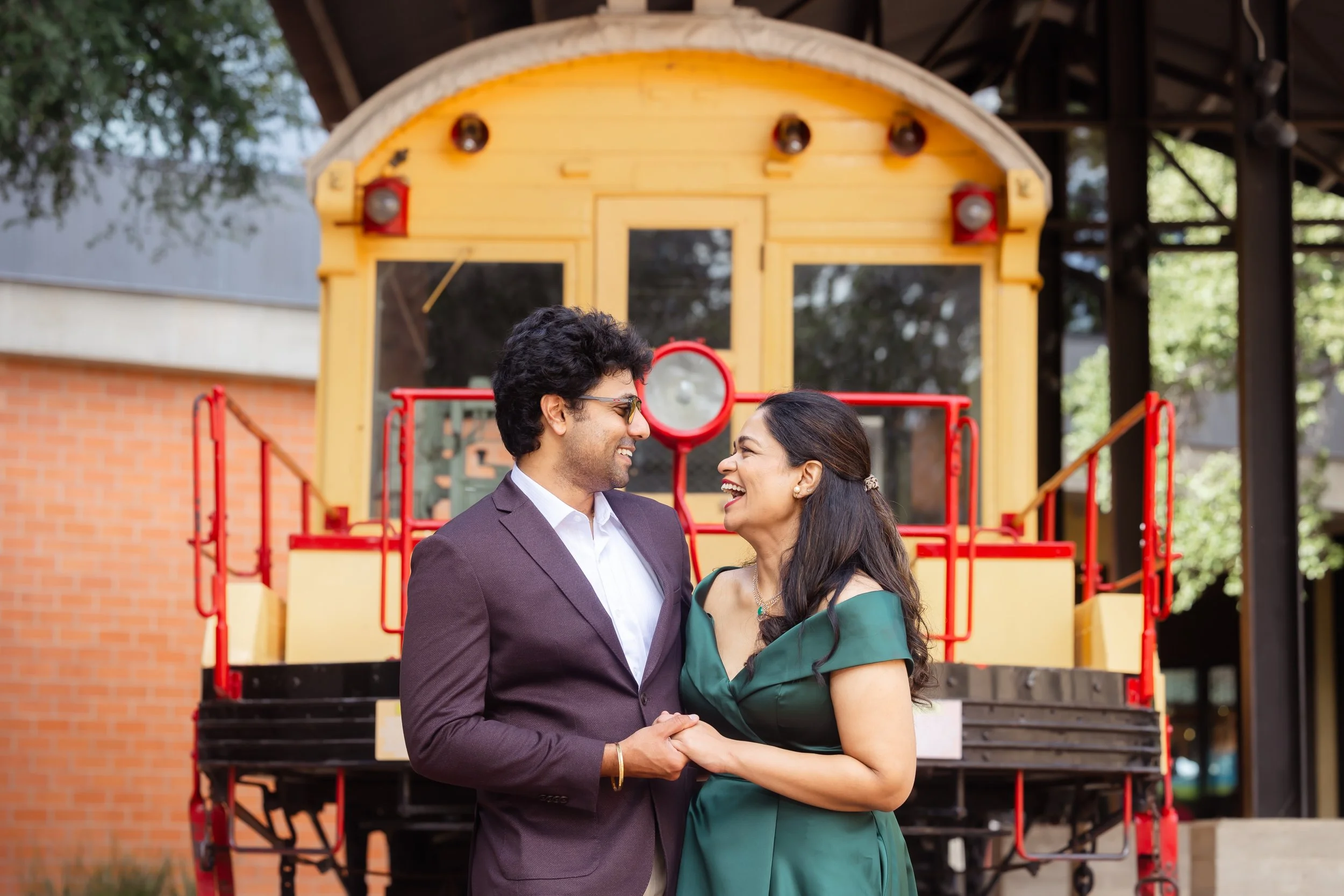 A happy couple holding hands and smiling at each other in front of a vintage yellow train with red accents.