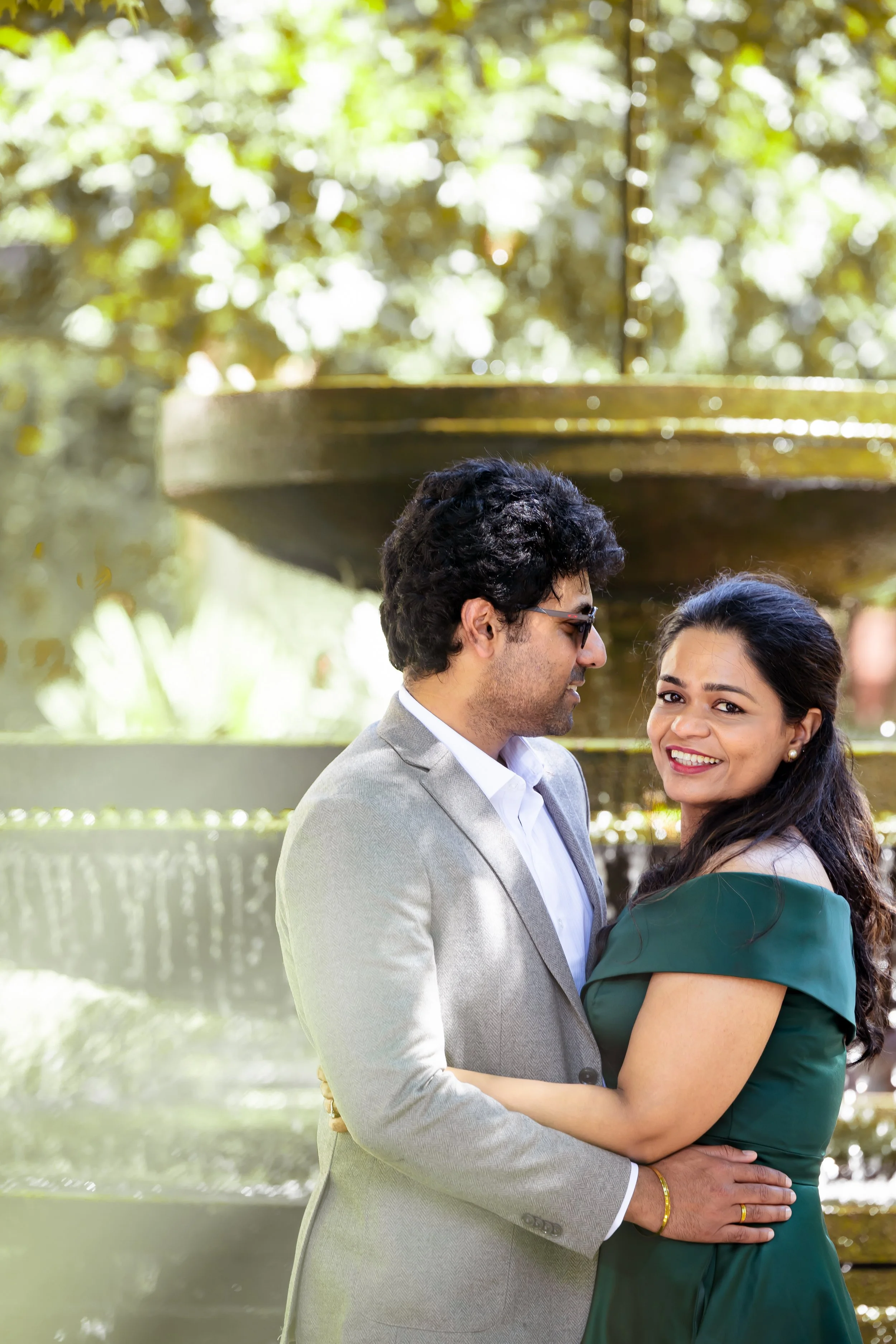 A couple stands close together outdoors, with the man wearing a light gray suit and the woman wearing a dark green dress, smiling at the camera near a fountain with sunlight filtering through trees in the background.