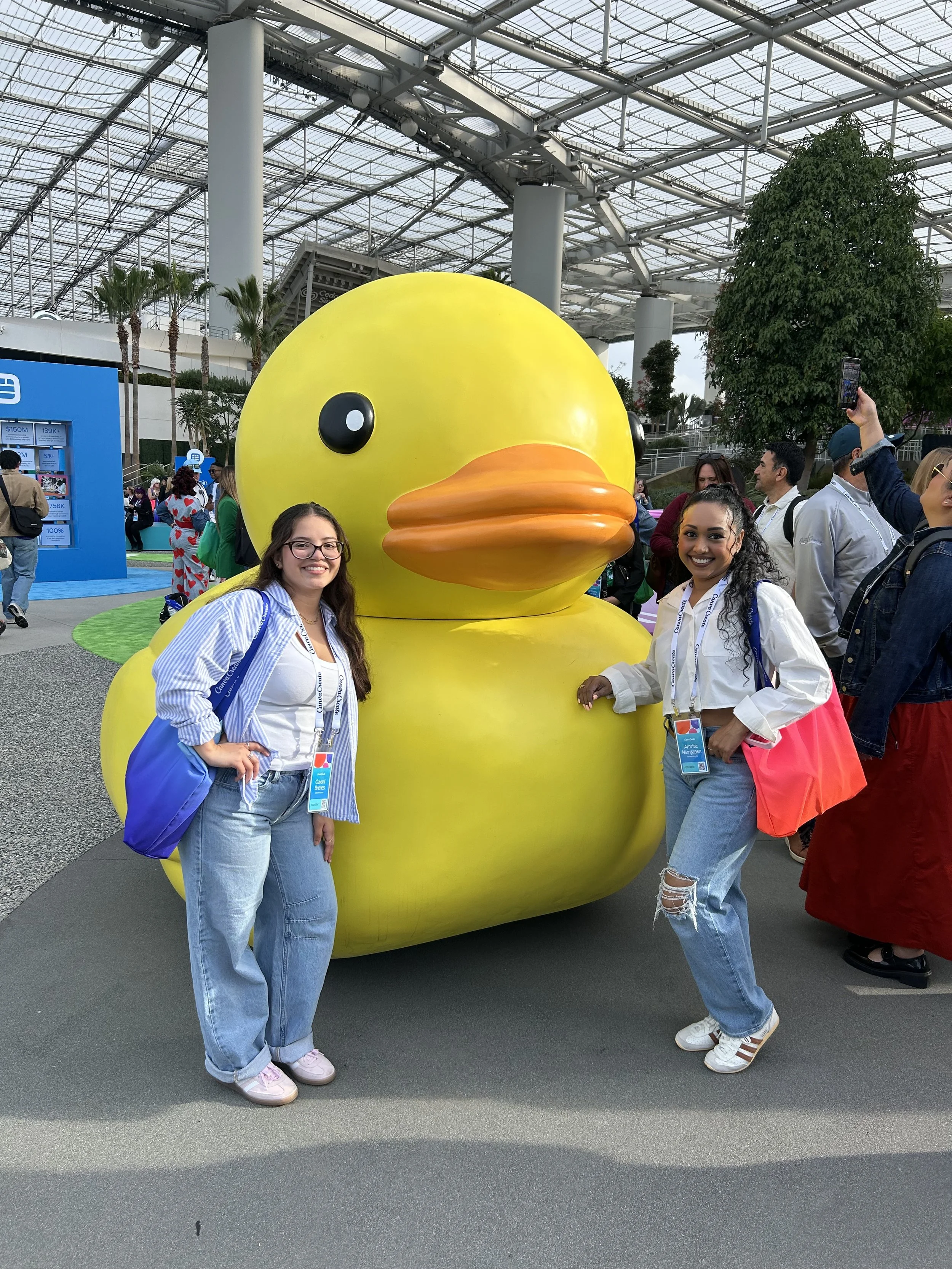 Two women standing next to a large yellow rubber duck at an indoor event with many people around.