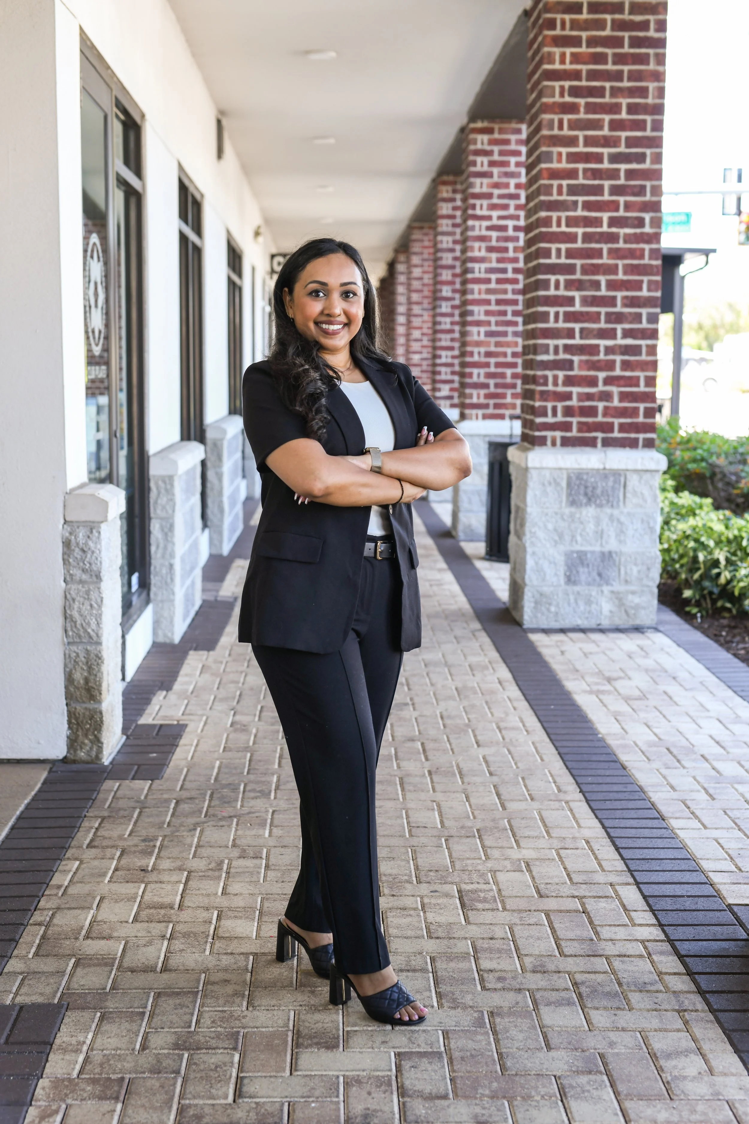 amrita in an black and white business casual outfit, 2/3rd postion, arms crossed, smiling, brick building background