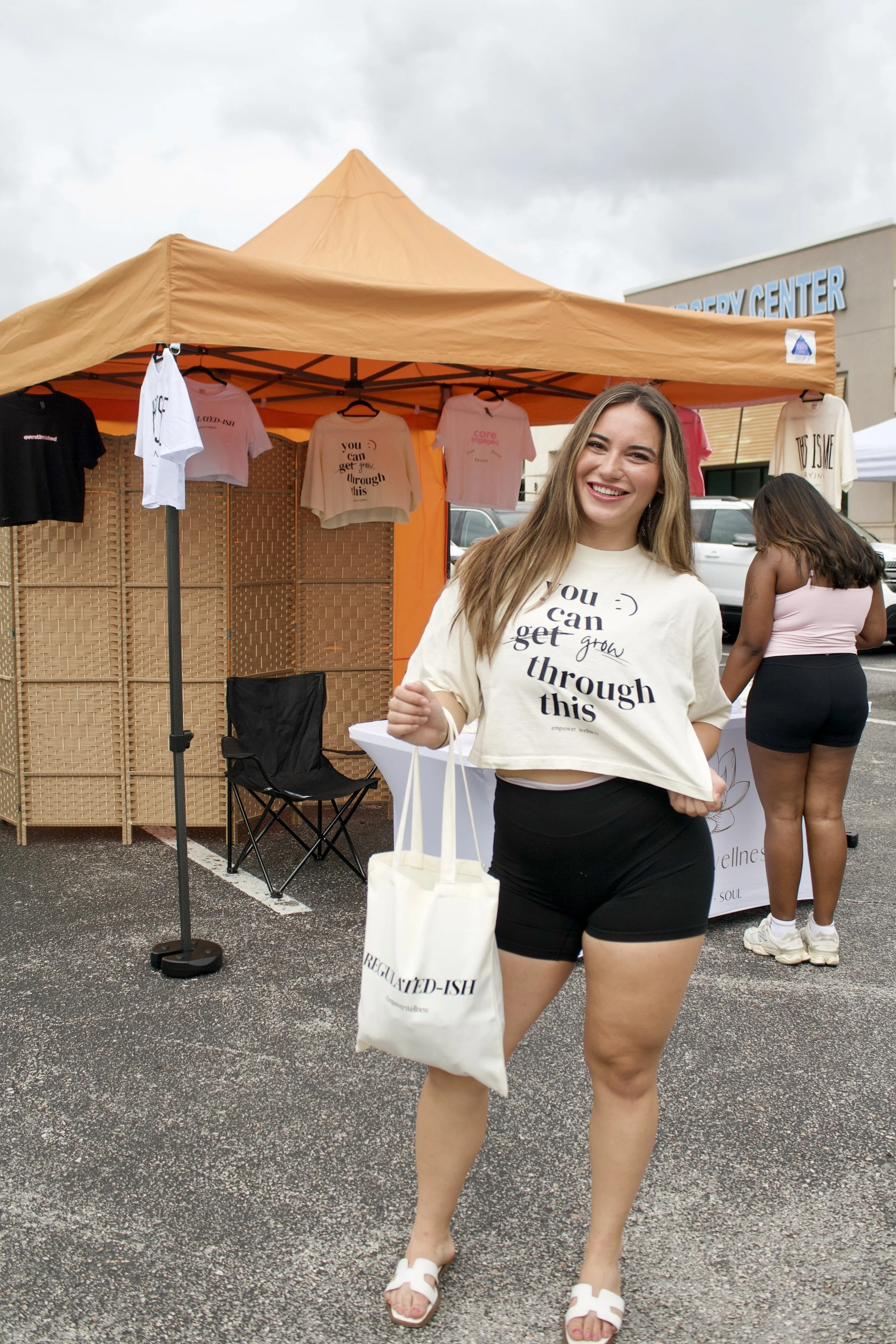 A smiling woman with long light brown hair, wearing a beige t-shirt with a motivational quote and black shorts, standing in front of a vendor booth at an outdoor event, holding a white bag.