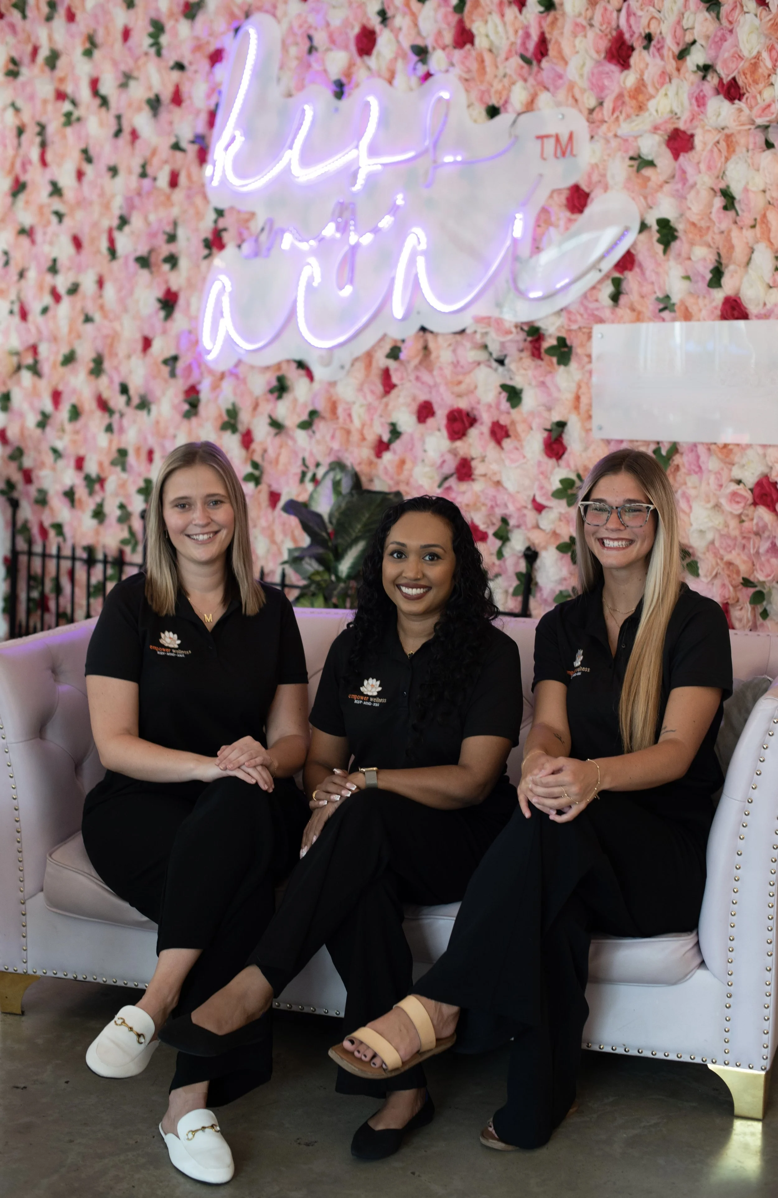 Three women sitting on a white couch, in black clothes, smiling. Rose covered wall behind them