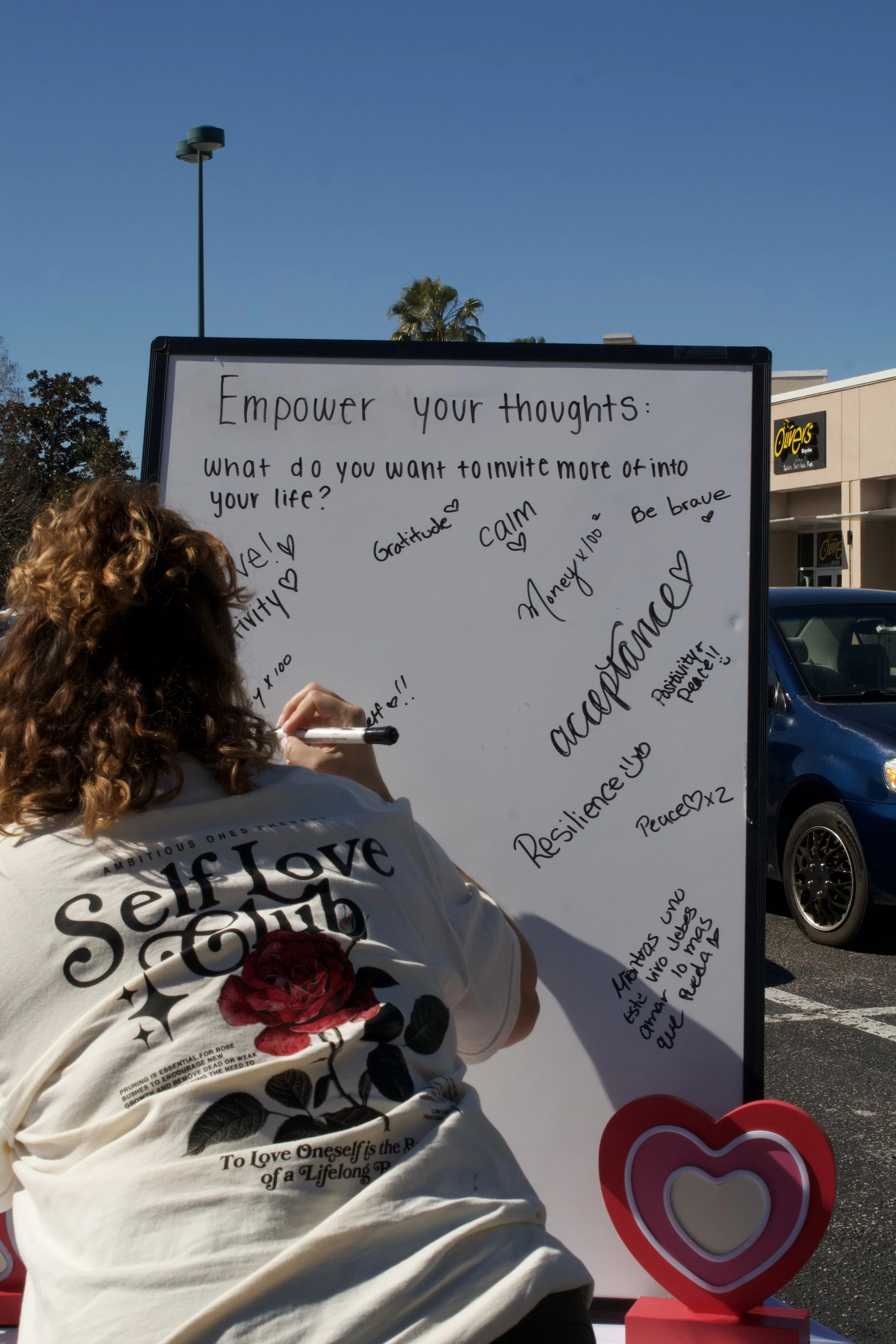 A person with curly hair wearing a white shirt with a floral design and the words "Self Love" written on it, writing on a whiteboard or poster outside in a parking lot with a blue sky and shopping center in the background, including a sign for Oliver's.