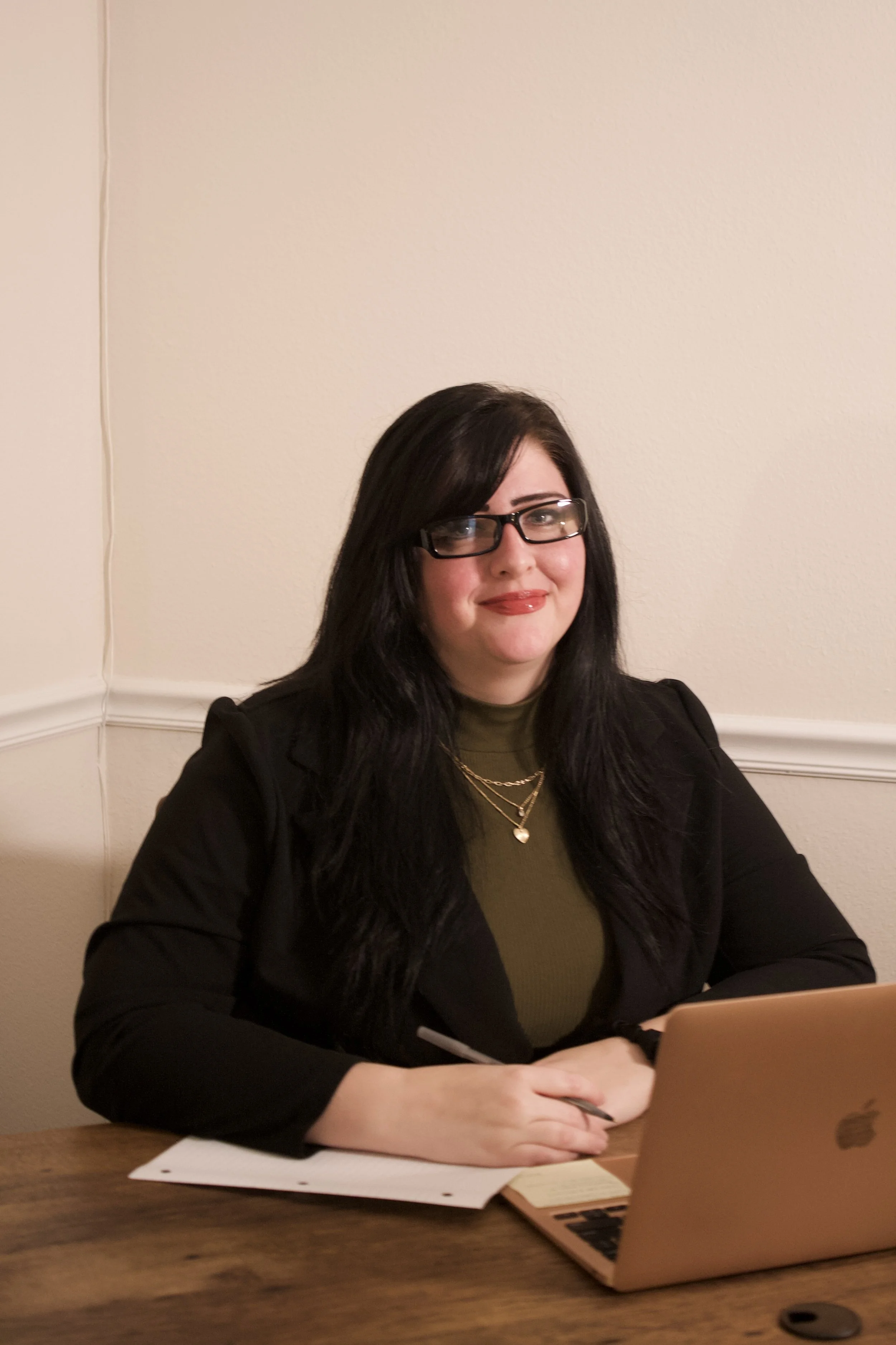 ashlie in a black and olive green business casual outfit, smiling at the camera, and sitting at a desk with a laptop, paper, and pen in hand