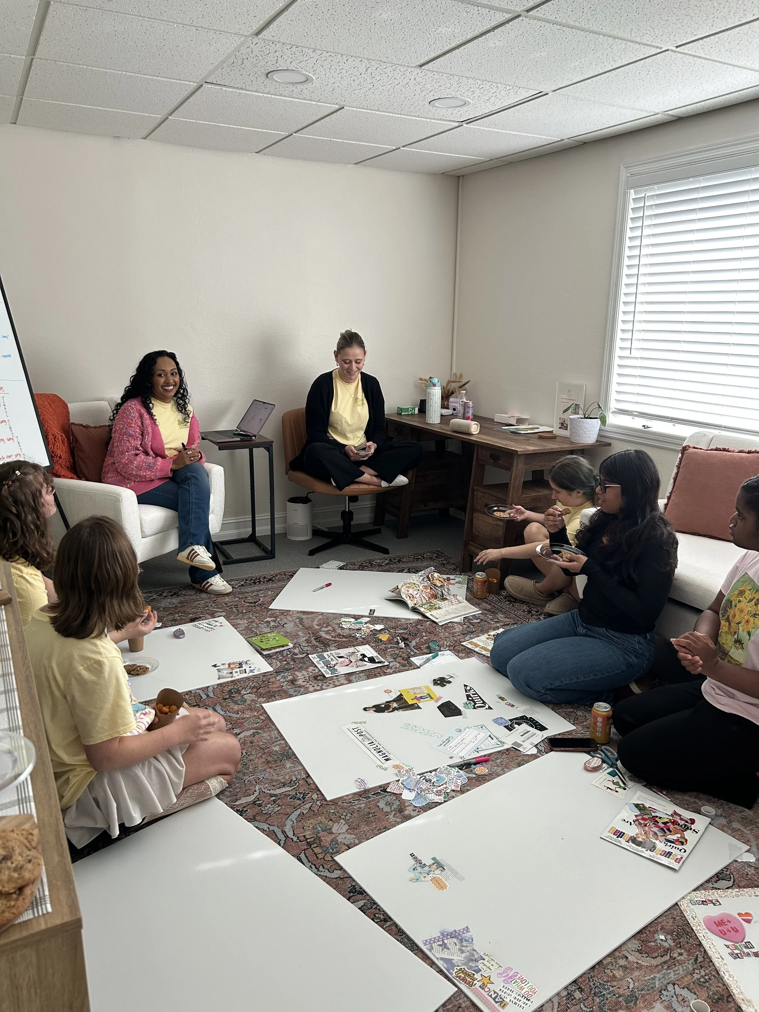 A group of children and two women sitting on the floor of a room, engaging in a craft or creative project with posters, magazines, and art supplies laid out in front of them.