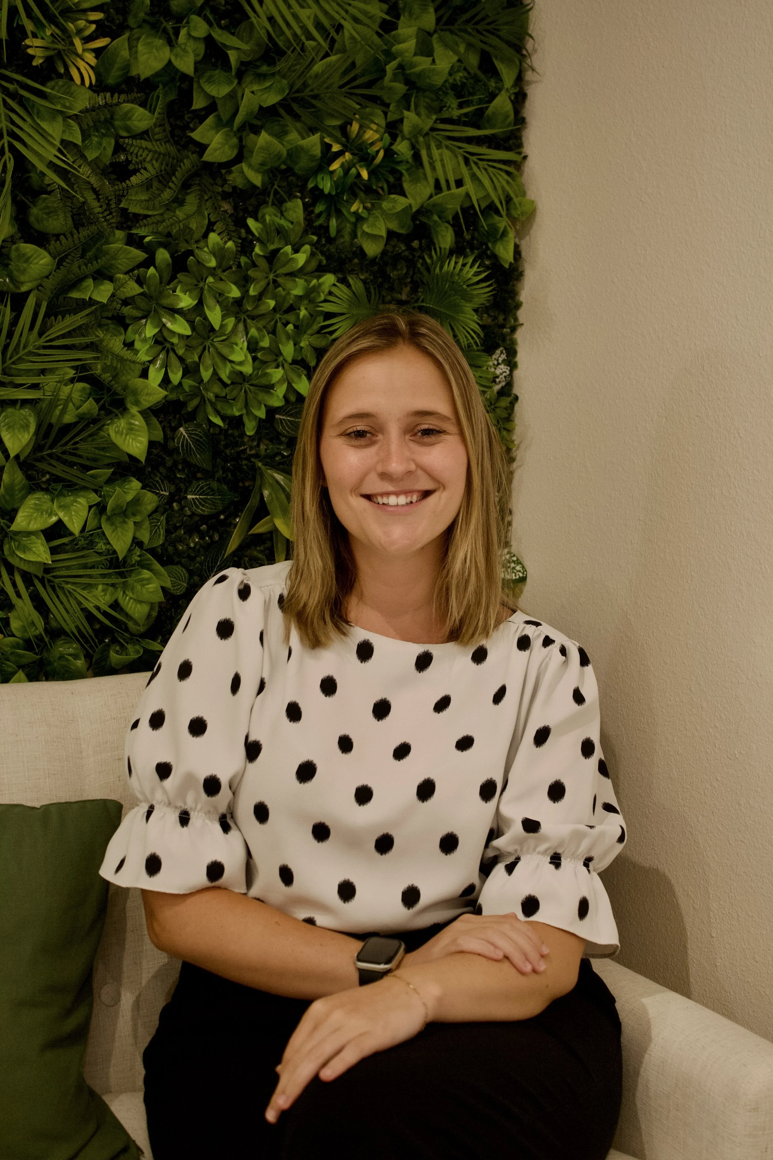 Morgan Arevalo sitting down in a black and white polka dot shirt, with a white wall and greenery wall in the back