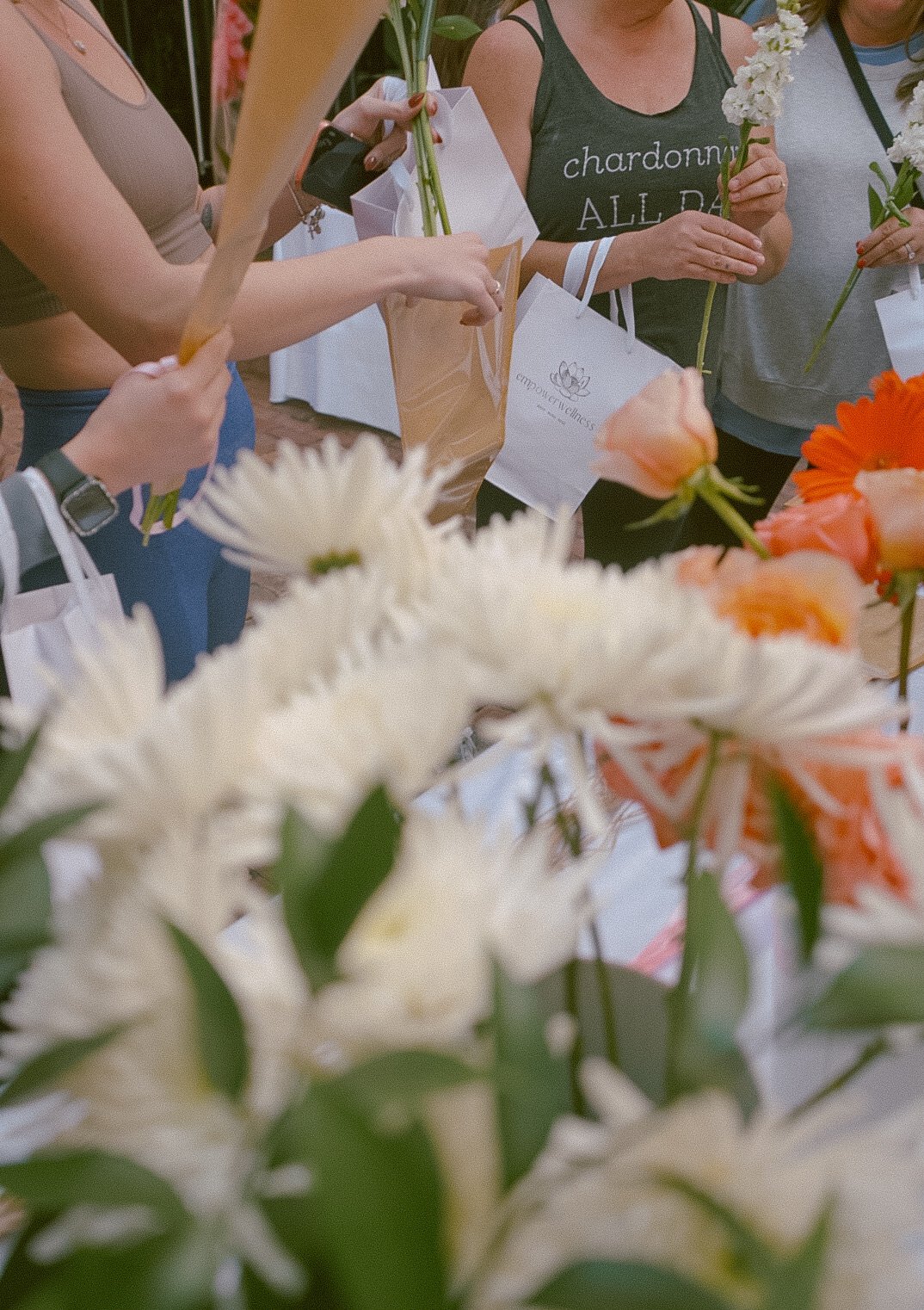 white daisies, peach roses, and orange daisies in majority of the photo with women in the background/top of photo, holding white empower wellness bags and flower bouquets