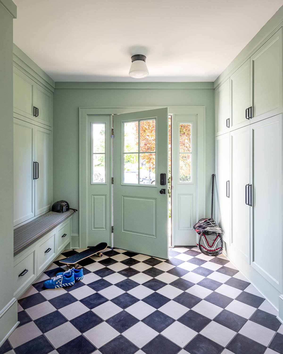 Entryway with pale green walls and checkered black and white floor tiles. A front door with glass panels opens to outside, showing trees with fall foliage. Shoes, a skateboard, a helmet, and sports equipment are in the space.