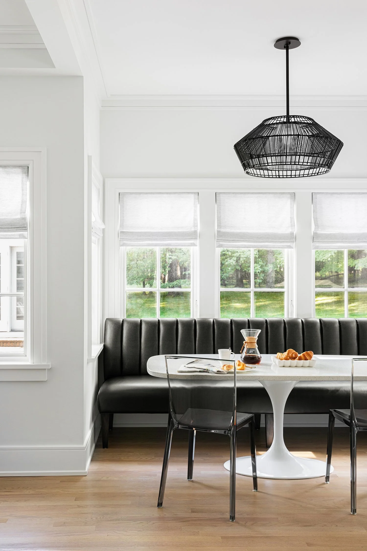 Modern dining area with a white round table, black chairs, and black bench seating. The table has a coffee pot, croissants, and a bowl of breadsticks. Large windows with white blinds let in natural light, and a black pendant light hangs from the ceil