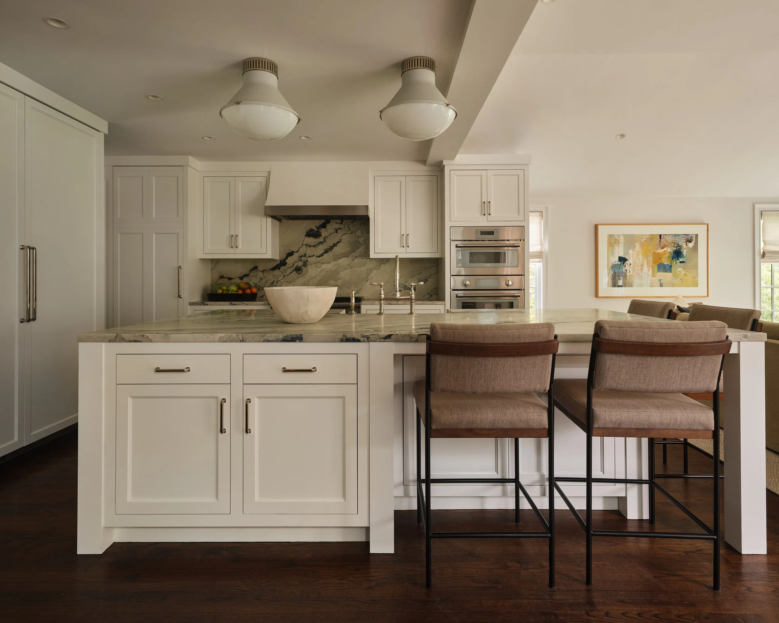 Modern kitchen with white cabinets, marble island, and barstools in front. There is a bowl of fruit on the countertop and a piece of abstract art on the wall. Ceiling lights are visible.