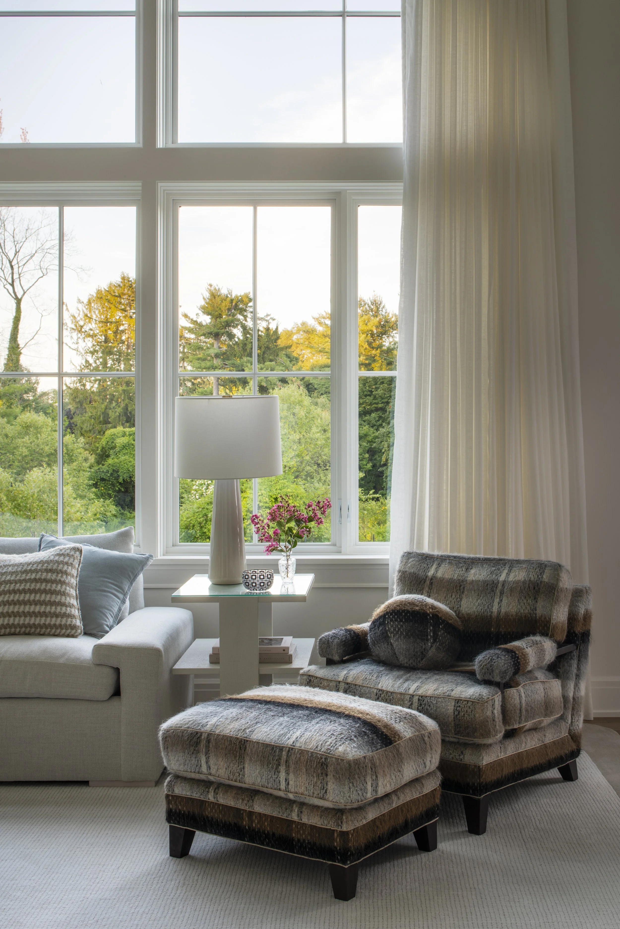 Living room with large window showing trees outside, beige sofa with striped and gray pillows, white side table with a white table lamp, pink flowers, and a plaid armchair with matching ottoman.