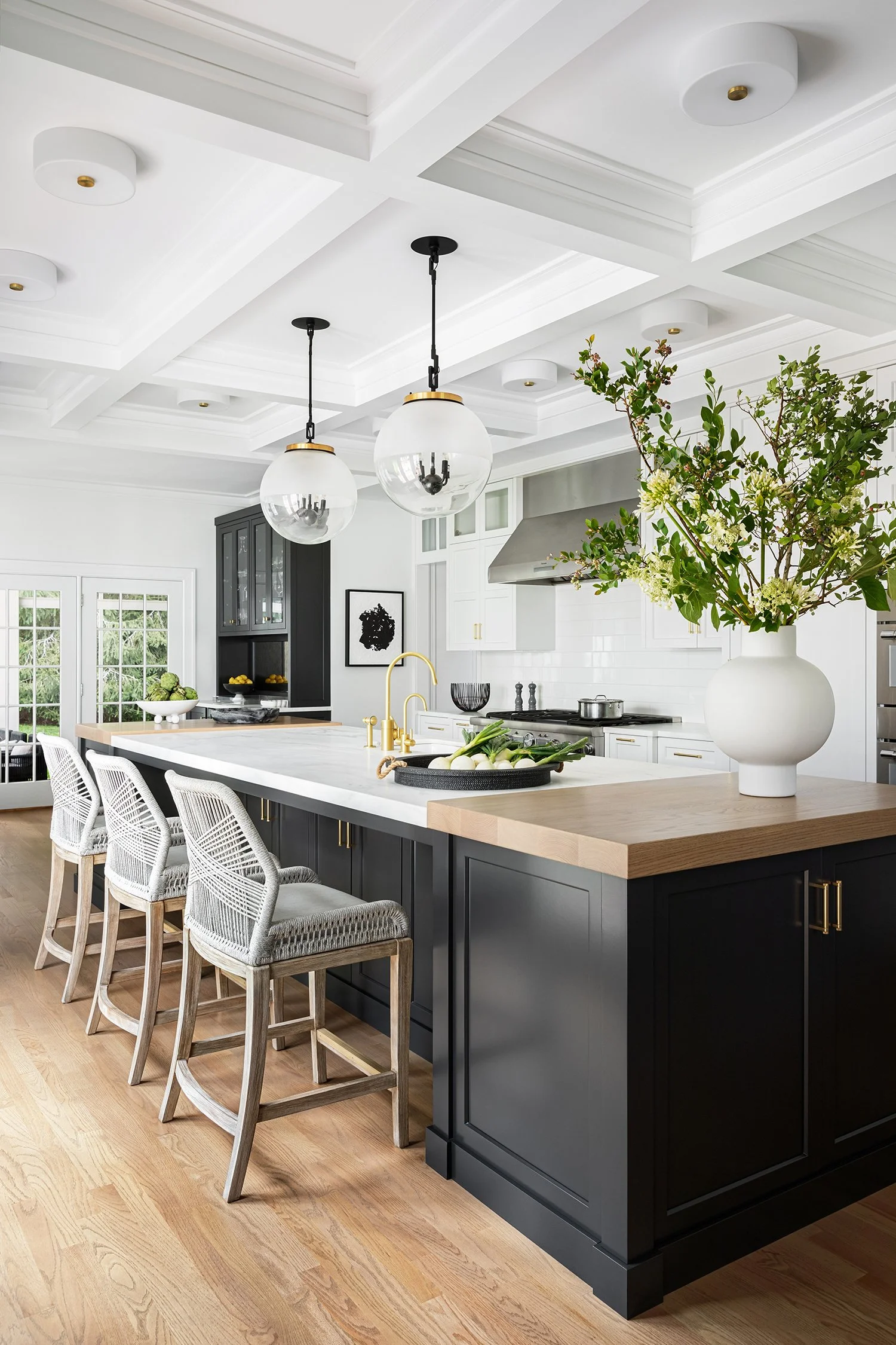Modern kitchen with black cabinetry, white countertops, gold fixtures, pendant lights, a large white vase with green and white flowers, and beige chairs around a kitchen island.
