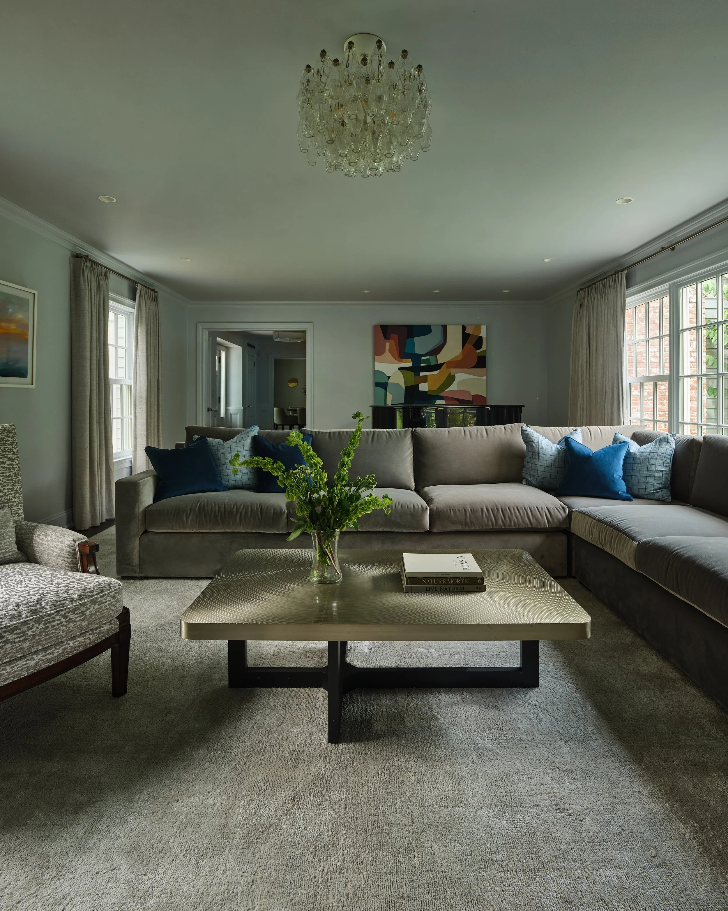 Living room with beige sectional sofa, blue pillows, black coffee table with a glass vase of green plants and stacked books, large window with cream curtains, abstract wall art, chandelier, and a doorway leading to another room.