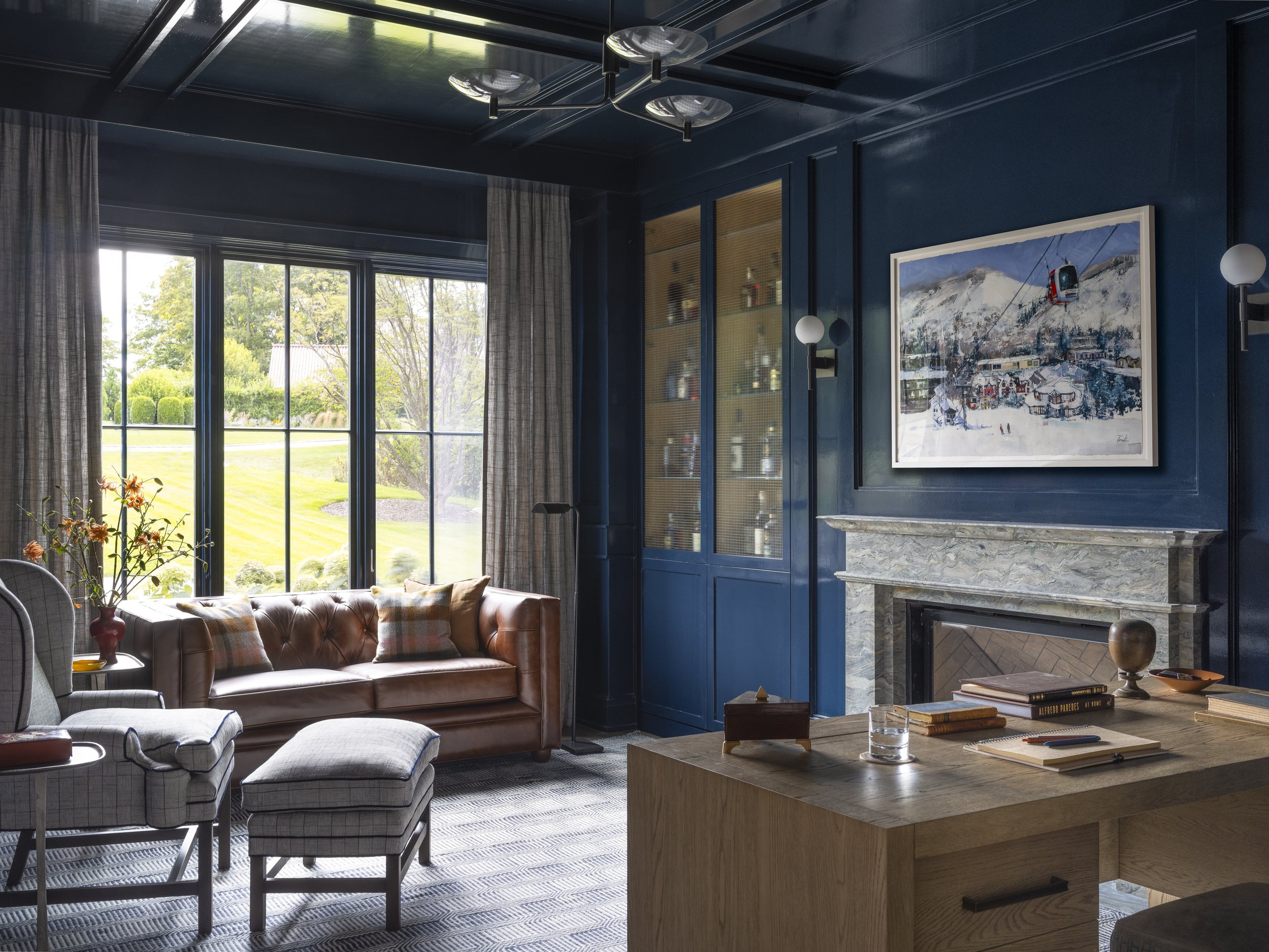 Living room with large windows, blue walls, a leather sofa, a checkered armchair, a marble fireplace, a wooden coffee table with books and glass, and a framed painting of snow-covered mountains and a cable car.