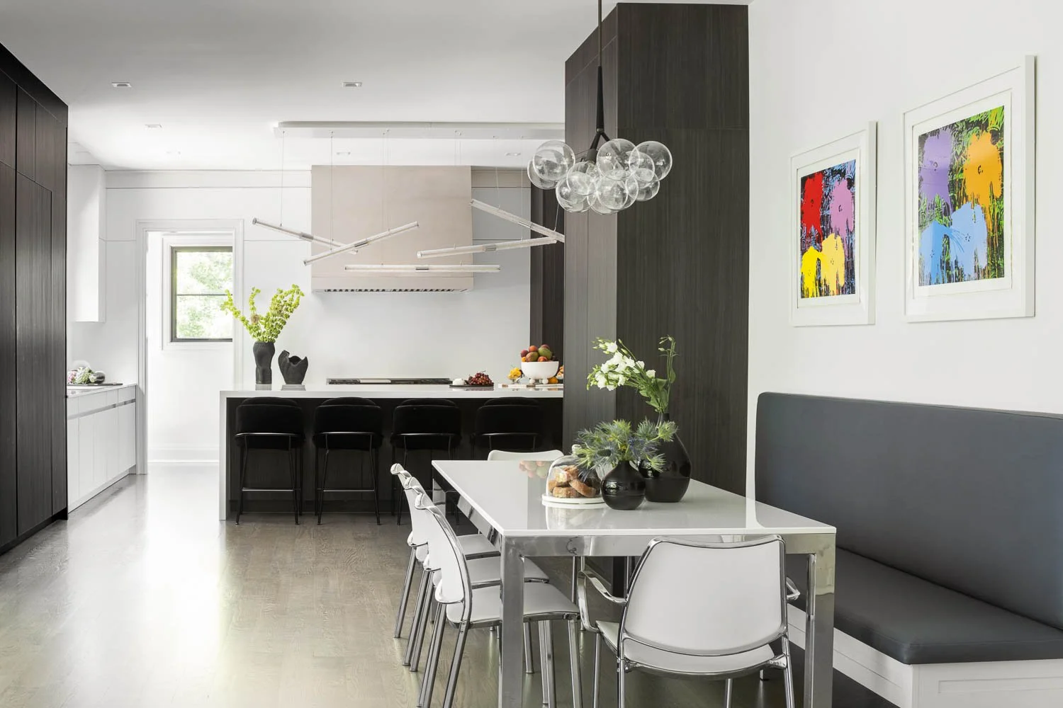 Modern kitchen and dining area with black, white, and gray decor, featuring a dining table with four chairs, a black bench, abstract colorful artwork on the wall, vases with green plants, and a kitchen island with bar stools.