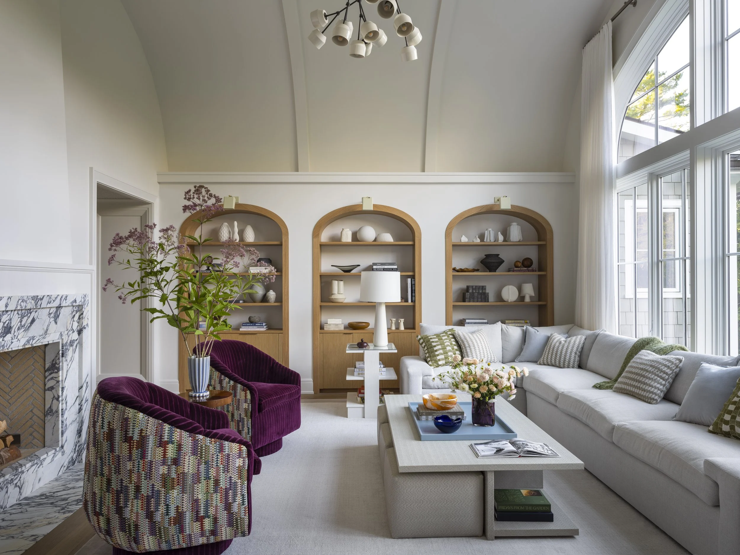 Living room with high arched ceiling, large windows letting in natural light, white sofa with patterned pillows, purple armchairs, built-in wooden shelves with decorative objects, a white coffee table with flowers and books, and a modern chandelier o