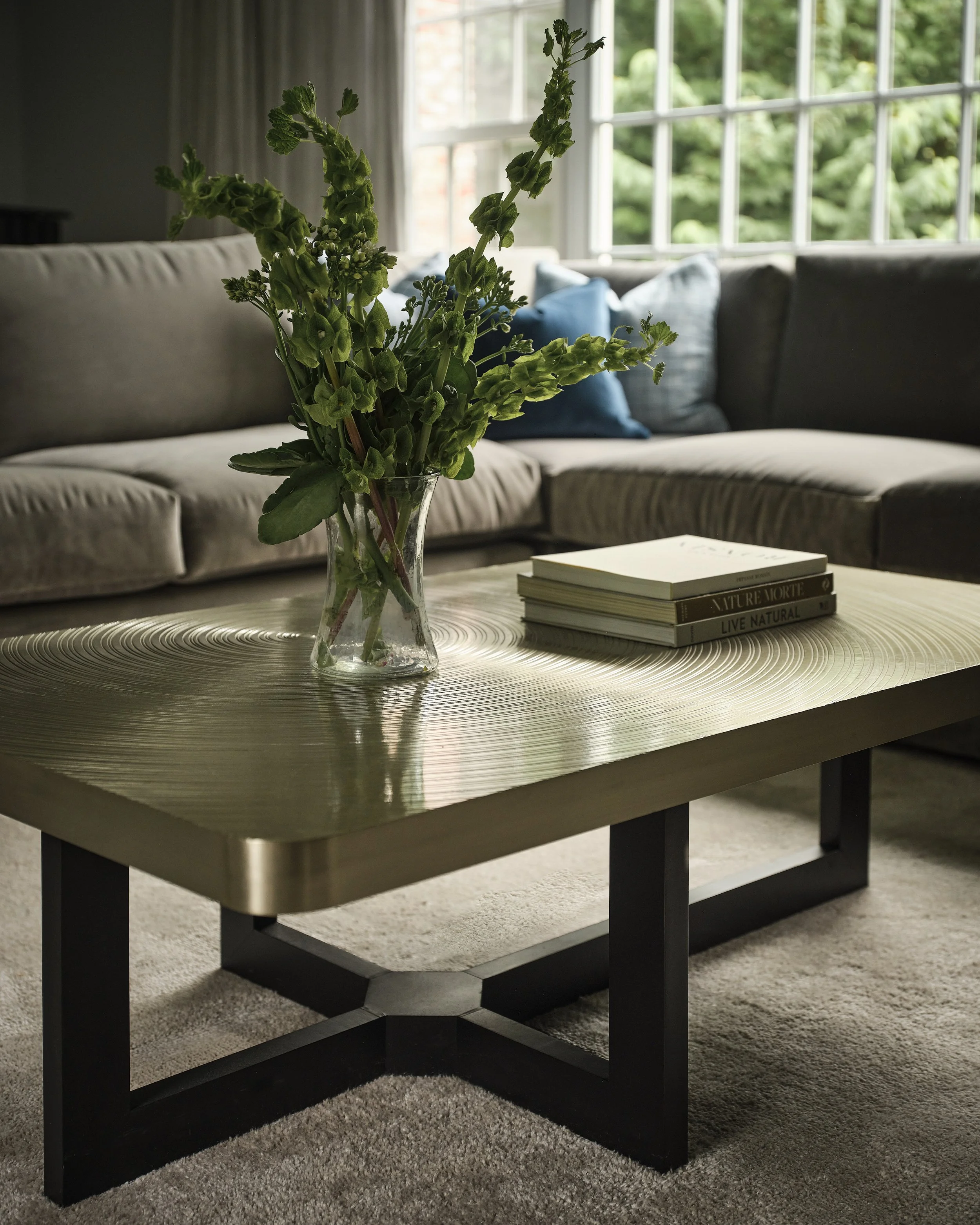 A living room with a beige and gray sofa, blue pillows, and a large window with white frames and curtains. In the foreground, a black and beige coffee table holds a glass vase with green leafy plants and a stack of books, one titled 'LIVE NATURAL'.