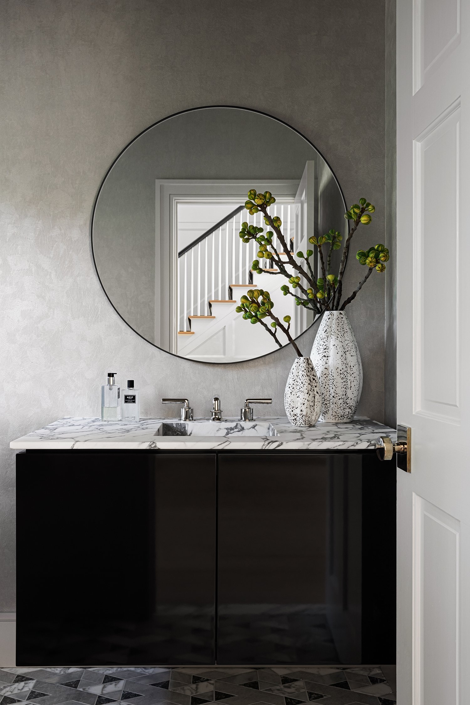 A bathroom vanity with a marble countertop, black cabinets, and modern fixtures. Two decorative vases with green plants and a mirror reflecting a staircase.