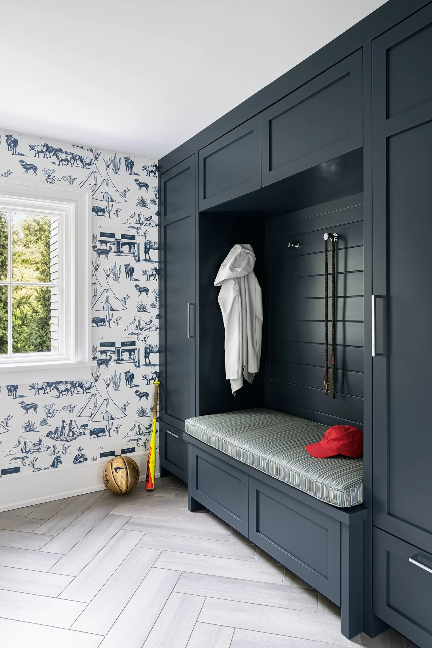 A built-in navy blue mudroom bench with a cushioned seat, a white jacket hanging on hooks, a red baseball cap on the cushion, a flag, a baseball, and a yellow mop leaning against the wall. The room has natural light coming from a window with white tr