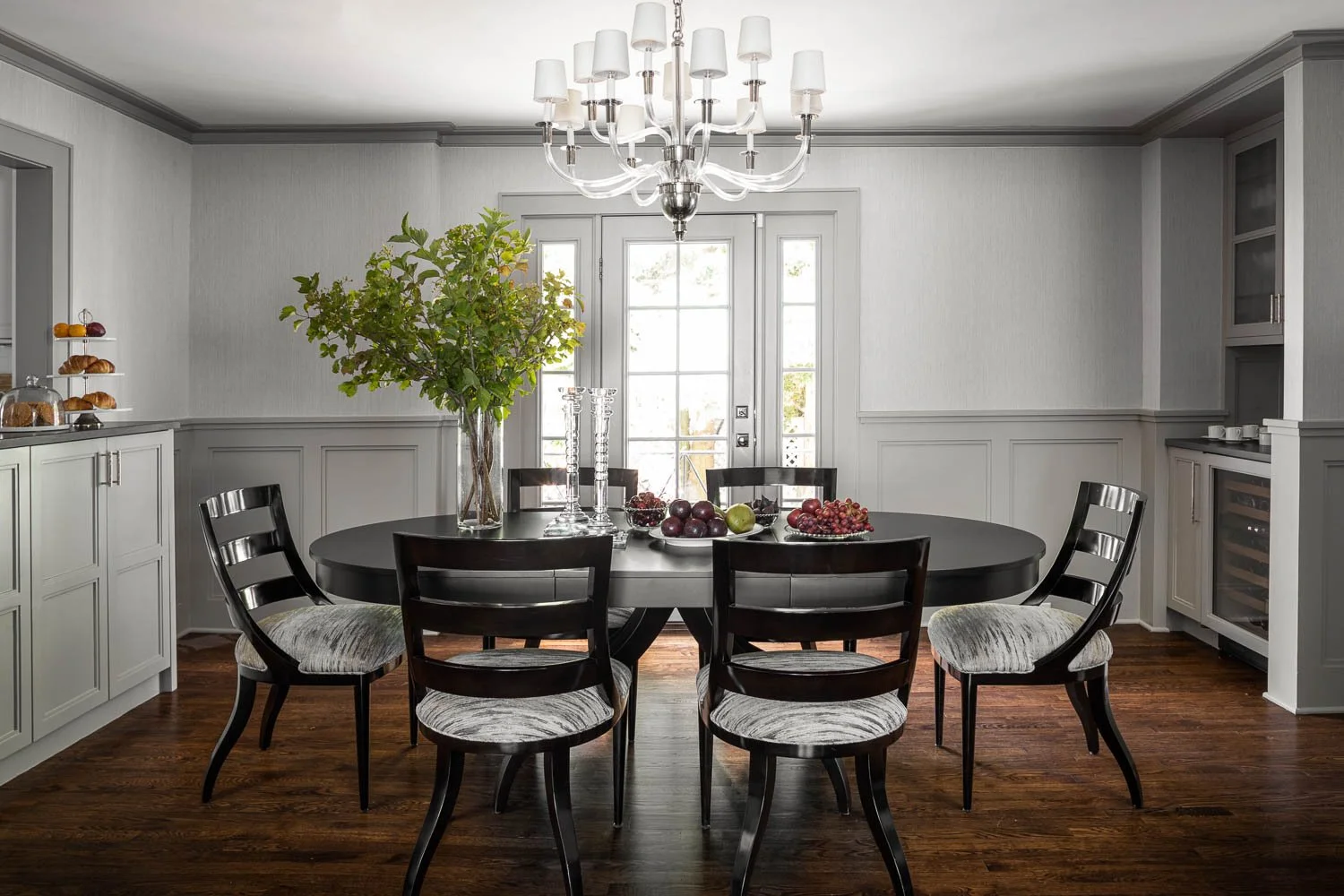 Dining room with a black oval table, six chairs with upholstered seats, large vase with green branches, fruits, and candles, and a chandelier hanging from the ceiling.