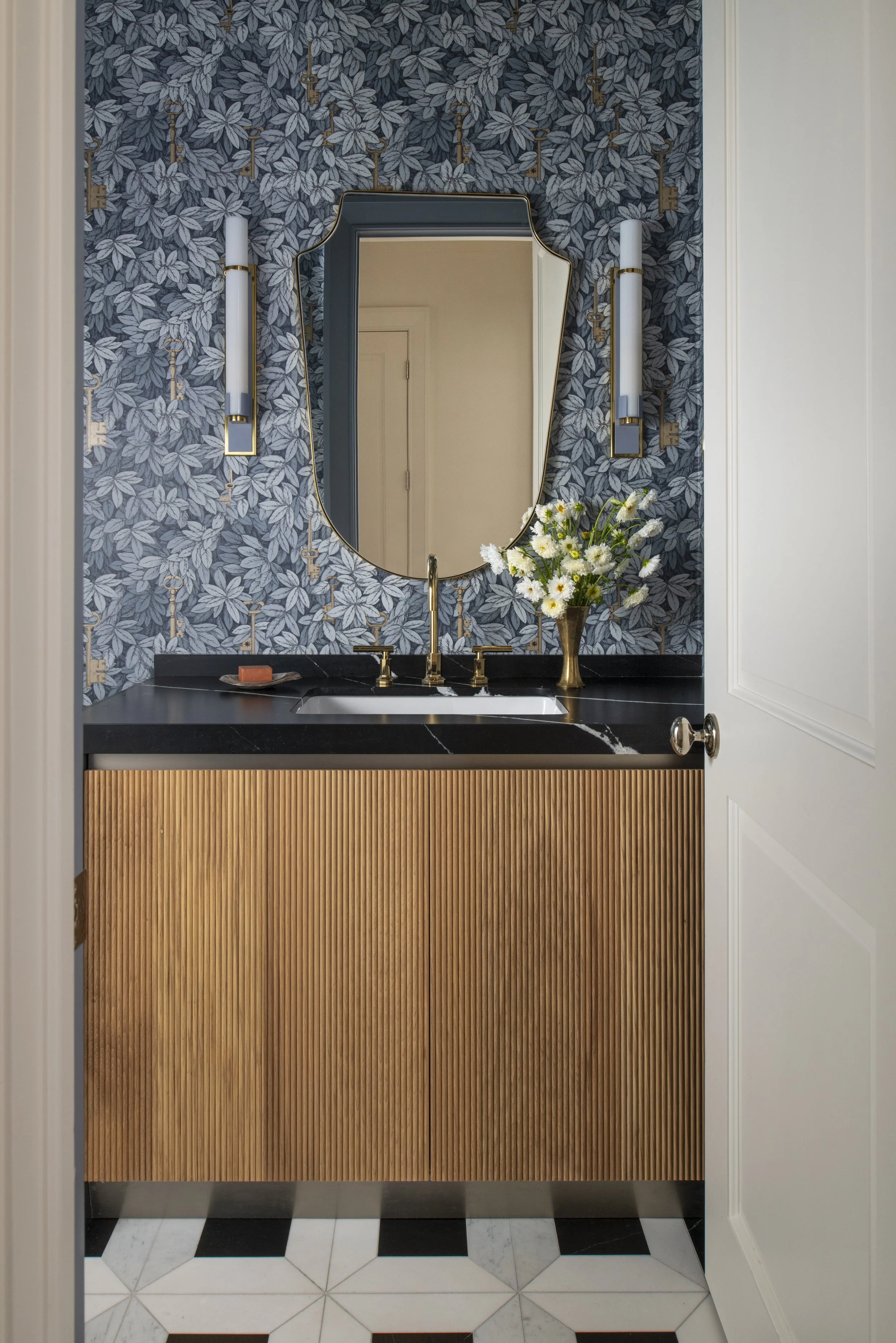 A bathroom vanity with a black countertop, gold faucet, and a wooden cabinet with vertical grooves. Above the vanity is a decorative mirror with a unique irregular shape, flanked by two modern wall sconces with gold accents and white cylindrical shad