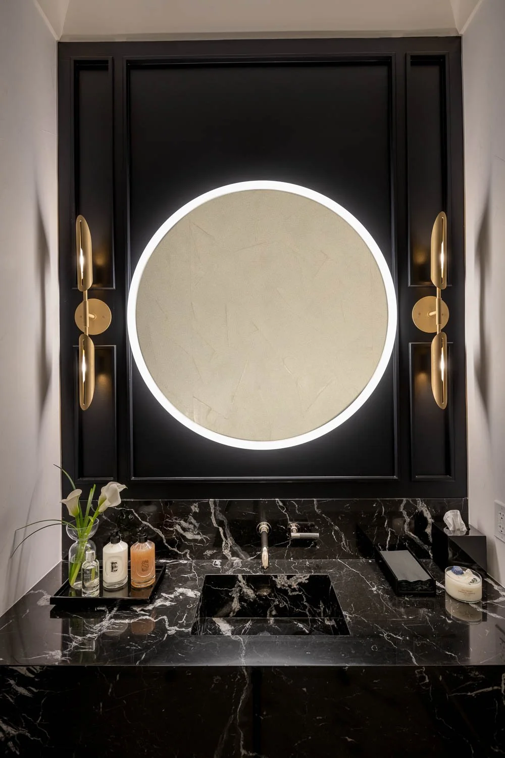 Modern bathroom vanity area with black marble countertop, round illuminated mirror, gold wall sconces, and small decorative items including a vase with white flowers and skincare bottles.