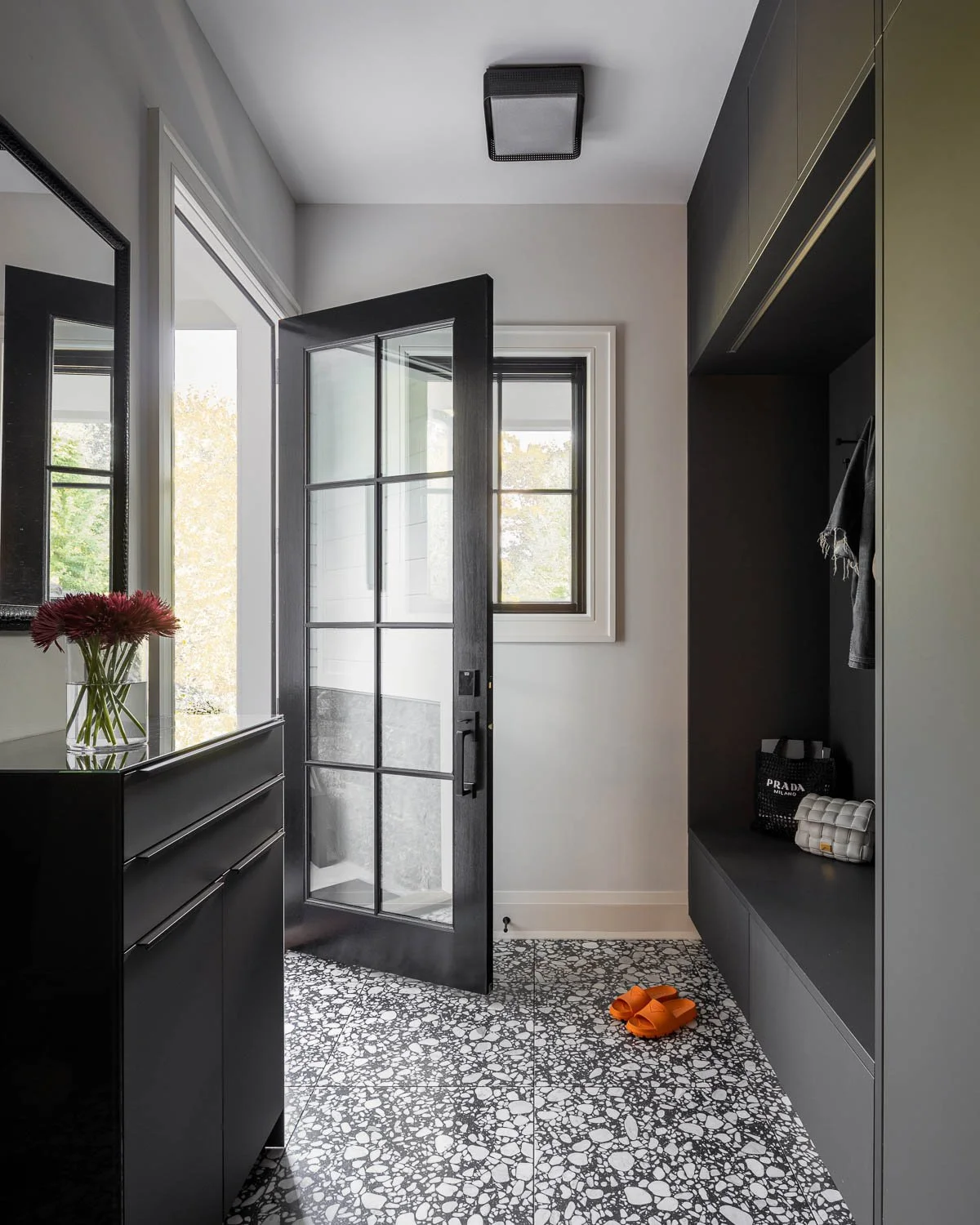 Modern entryway with black front door with glass panes, black and white terrazzo flooring, a black dresser with a vase of flowers, and black storage unit with closet items. Natural light coming through windows.