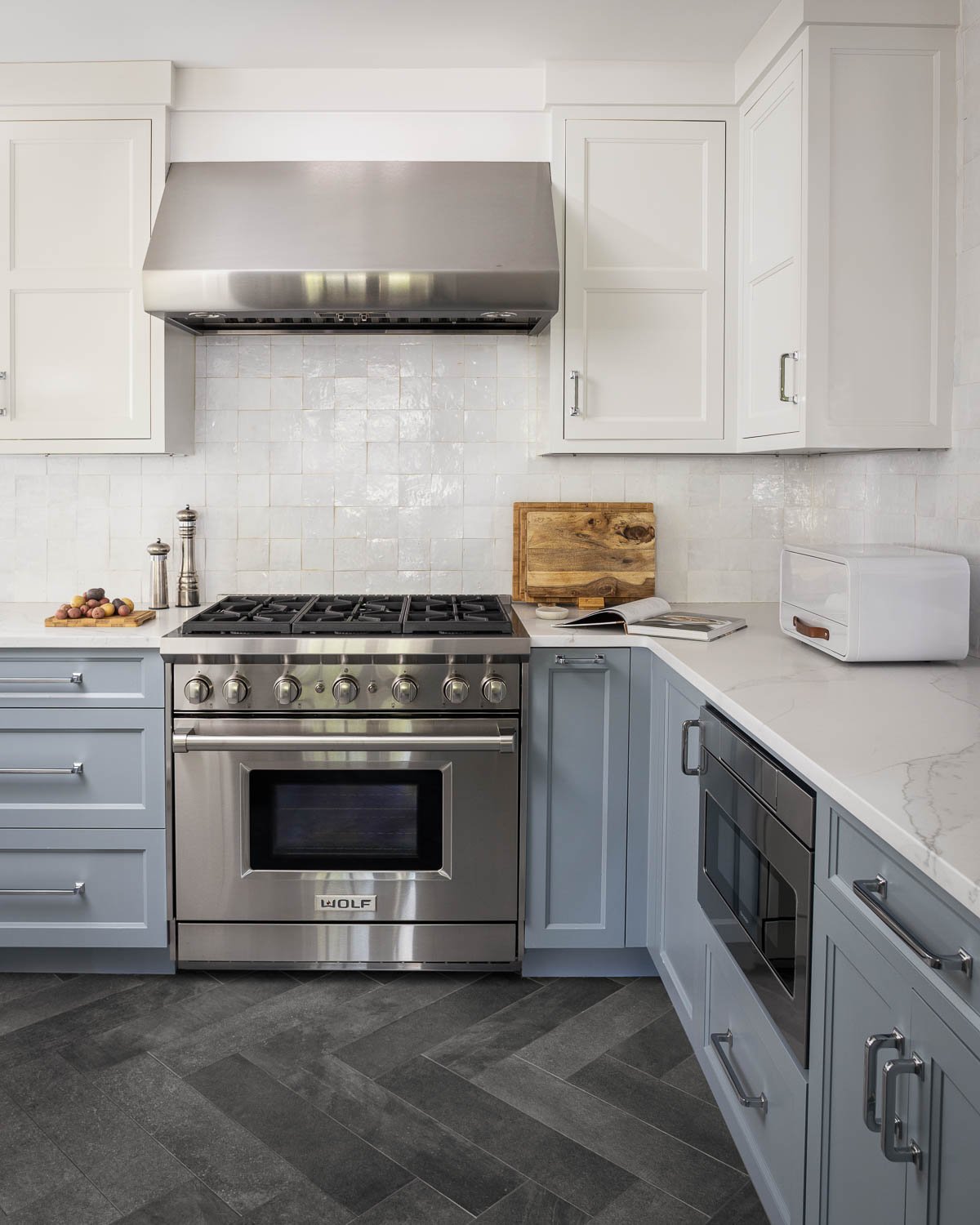 Modern kitchen with white upper cabinets, gray lower cabinets, stainless steel stove, white countertop, cutting board, open cookbook, white toaster oven, and gray tile flooring.