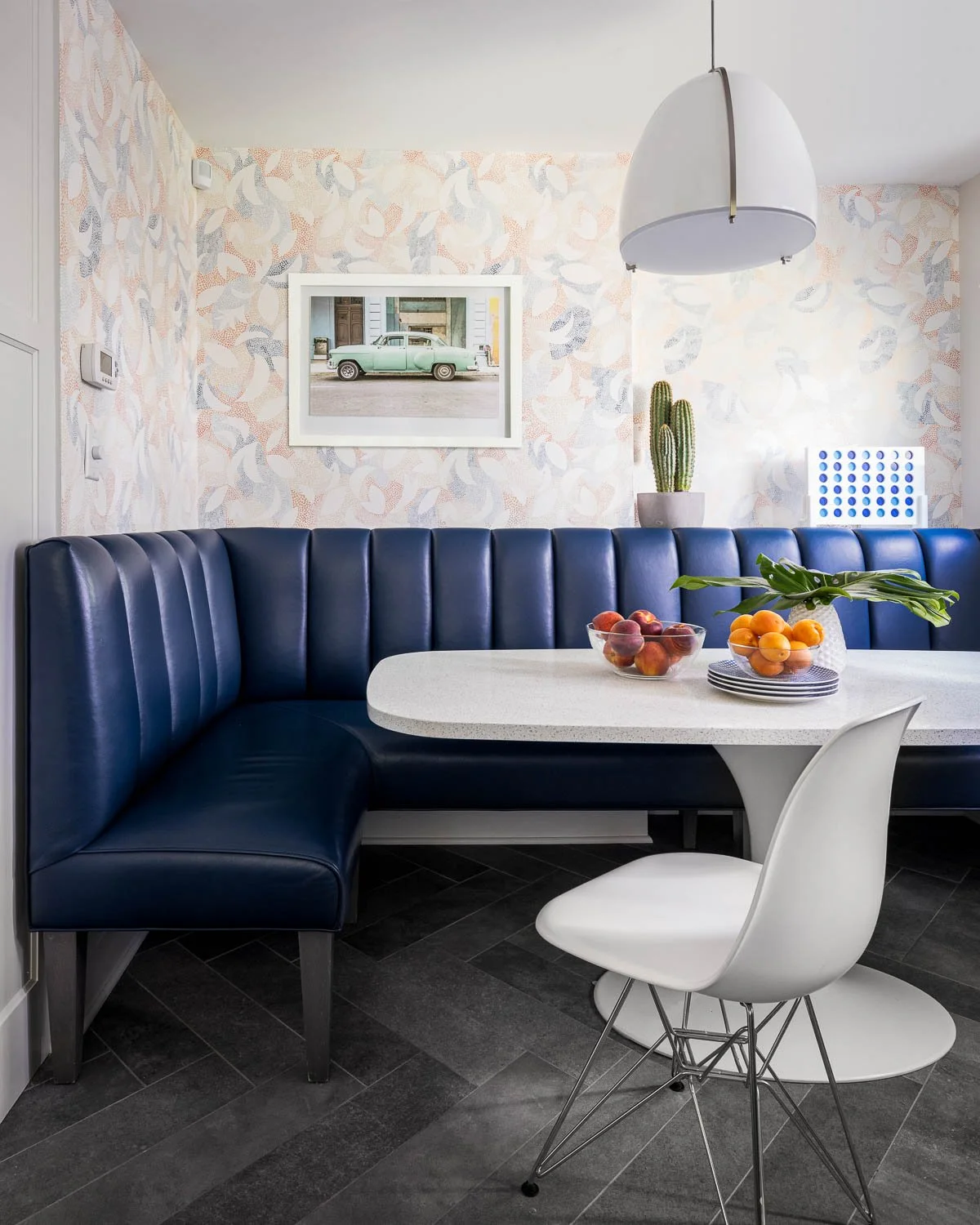 Modern dining area with a white oval table, a white chair, a navy blue curved booth, bowls of peaches and purple fruit, a green leaf arrangement, a cactus in a gray pot, a framed photo of a vintage green car, patterned wallpaper, a cactus, a white pe