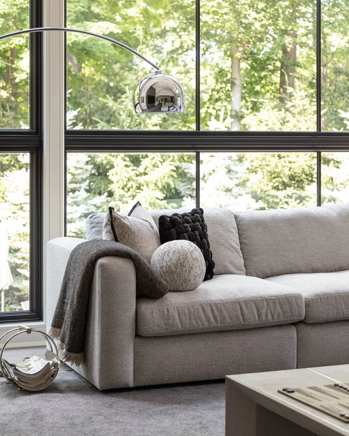 Living room with a gray sofa decorated with beige, black, and patterned pillows, a throw blanket, a round decorative object, and a modern silver arc floor lamp, set against large windows showing green trees outside.