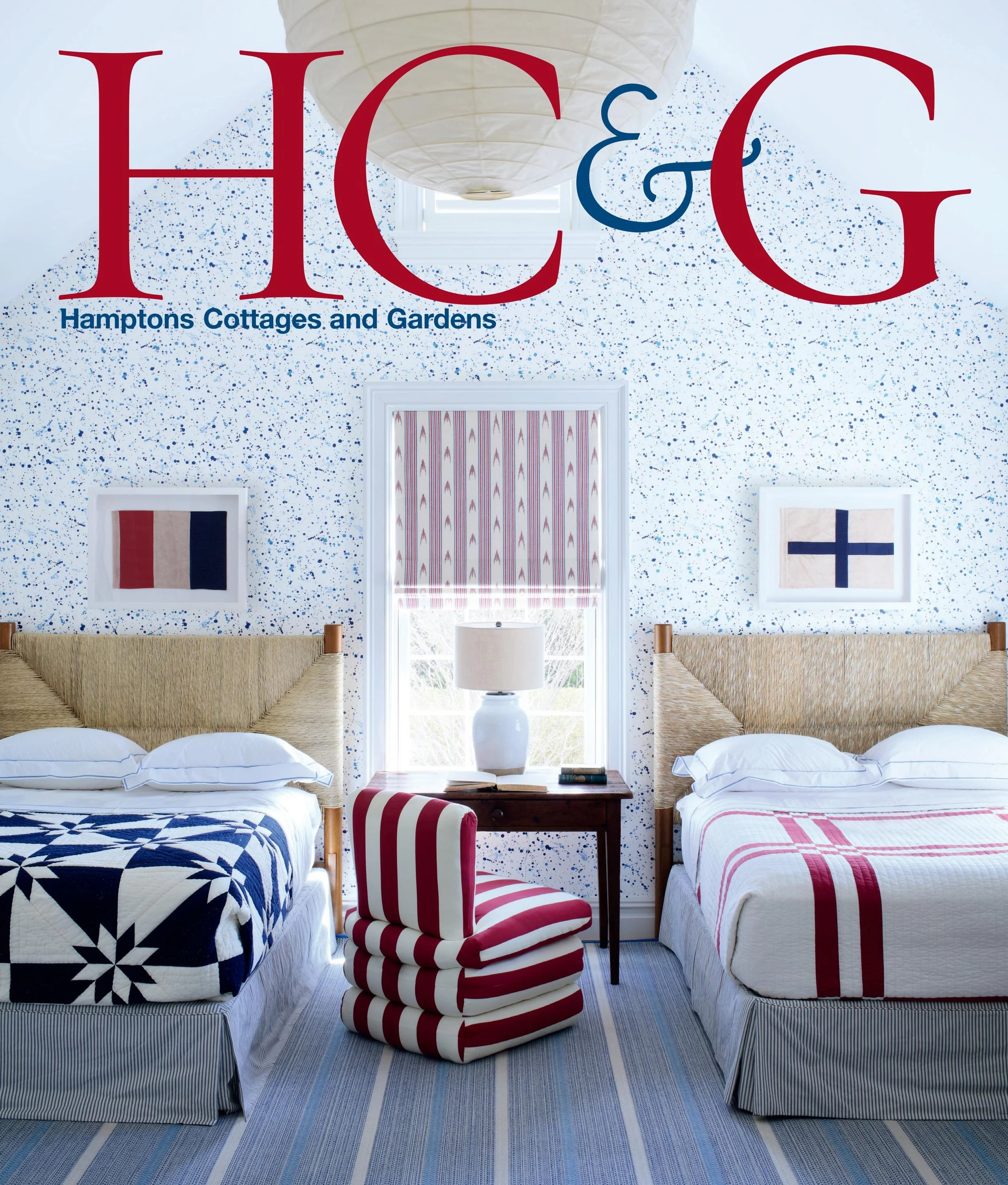 Two beds in a bedroom with white bed skirts, blue and white bedding, and American flag-themed pillows. Between the beds is a wooden nightstand with a white lamp and a book. The walls are light with blue speckles, and three square flags (French, American, and Finnish) are hanging above the beds. A window with a striped red, white, and blue curtain is at the back, and a striped blue rug covers the floor.