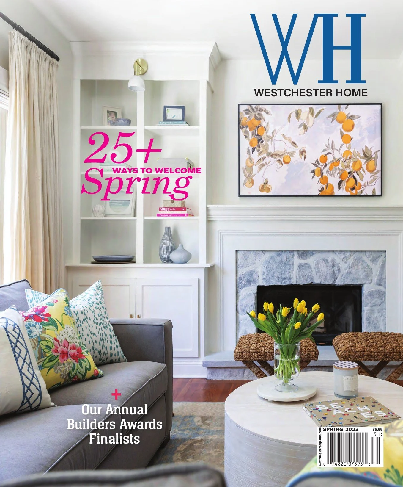 Living room with a white fireplace, colorful pillows on a gray couch, a white coffee table with a vase of yellow tulips, and wall art featuring orange fruit branches. The magazine cover headlines mention spring ideas and the annual builders awards finalists.