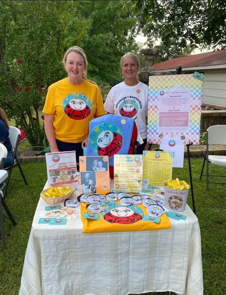 Two women standing behind a table displaying ladybug-themed items and informational materials at an outdoor event.