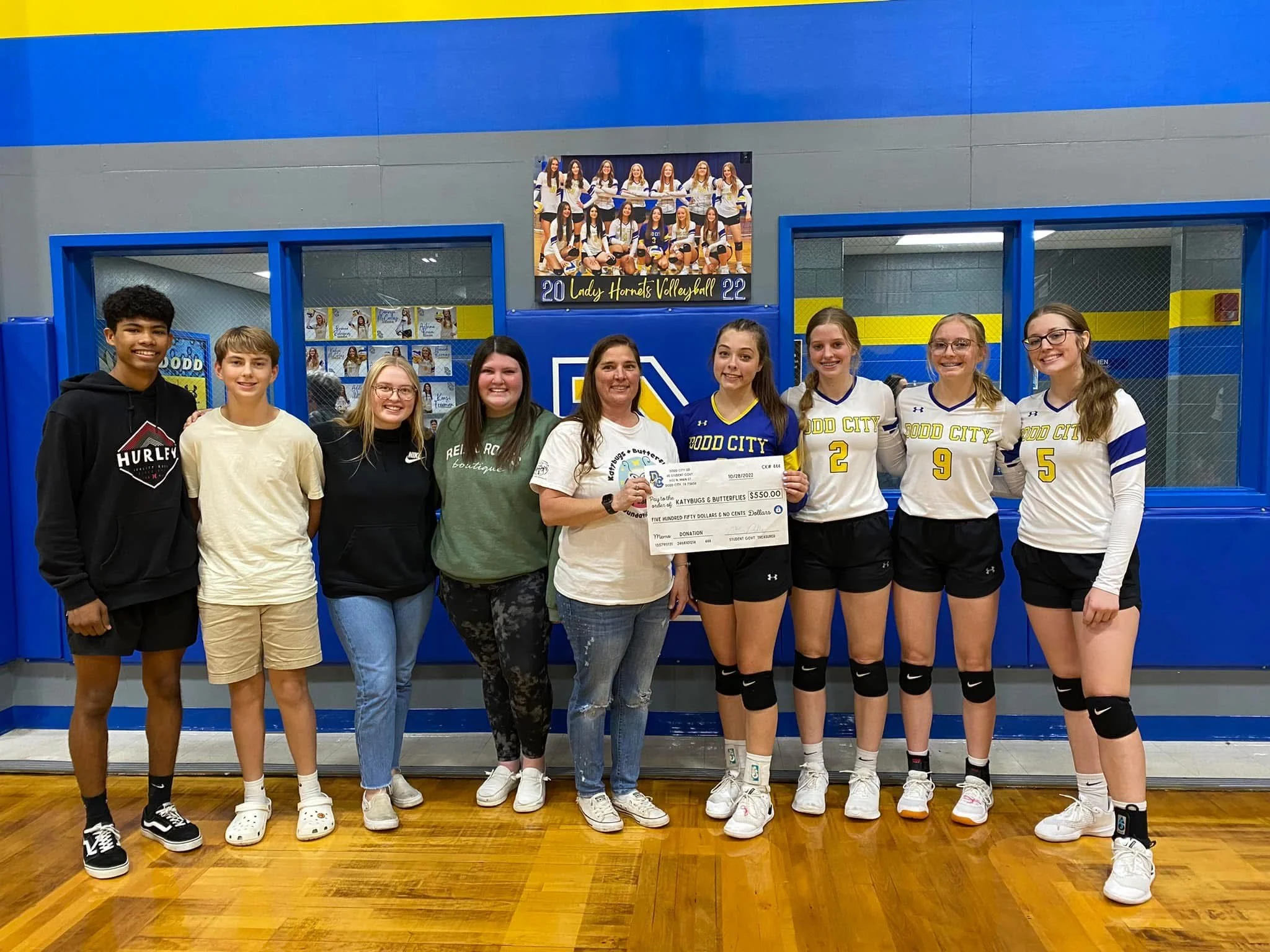 Group of young women and girls in volleyball uniforms celebrating with a large check in a gymnasium.