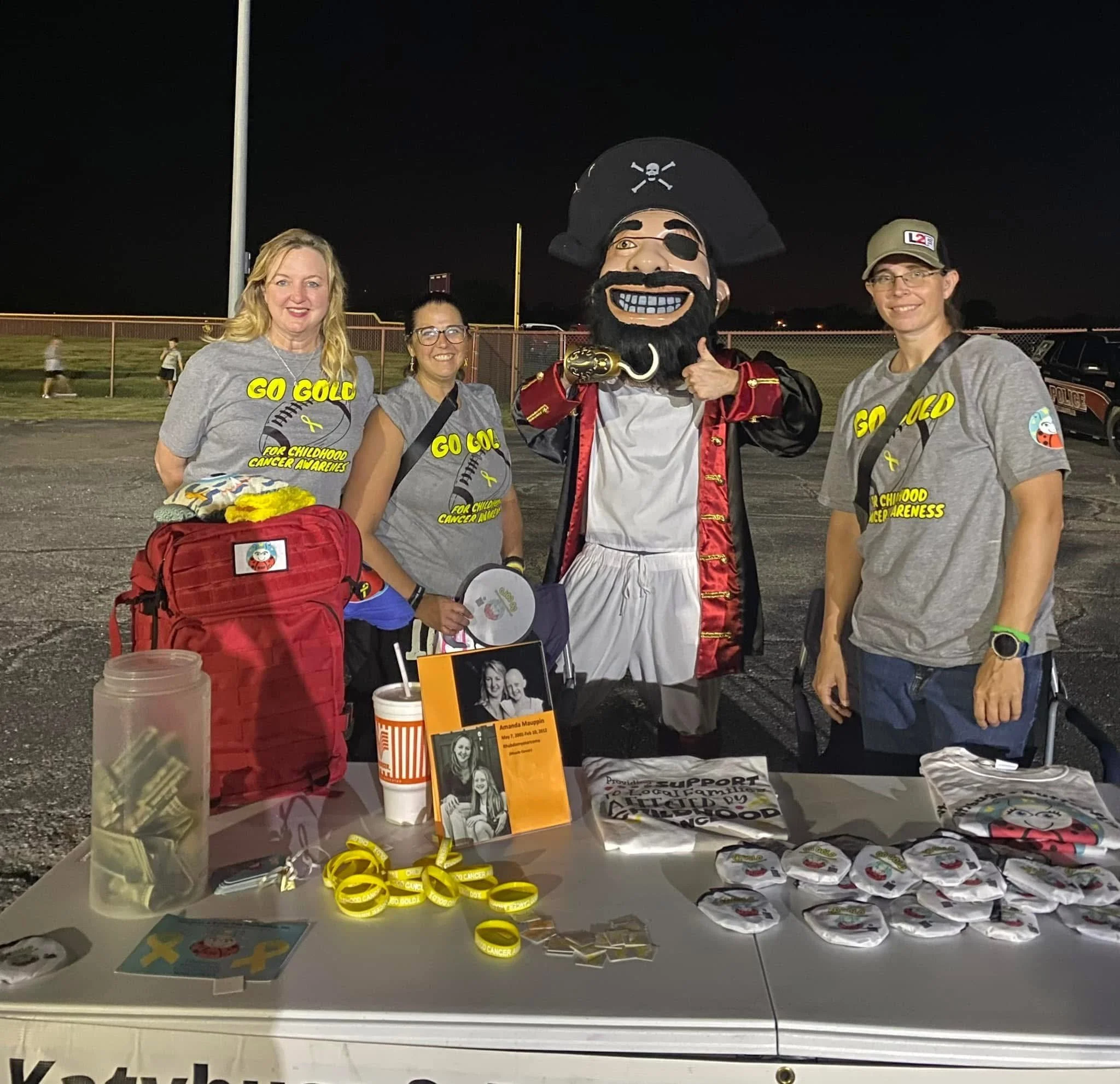 Four women and a person in a pirate costume standing behind a table at an outdoor event during evening. The women wear gray t-shirts with yellow text and ribbons for childhood cancer awareness. The table displays yellow bracelets, buttons, pamphlets,