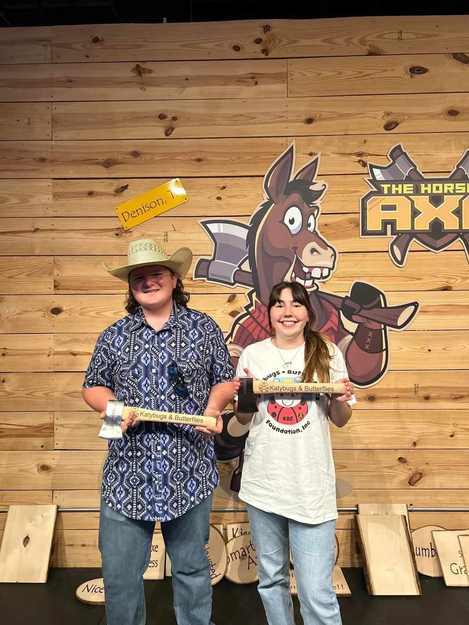 Two children standing in front of a wooden wall with a cartoon horse logo, holding wooden awards labeled "Katybugs & Butterflies." The boy on the left wears a cowboy hat and a patterned shirt, while the girl on the right wears a white T-shirt featuri