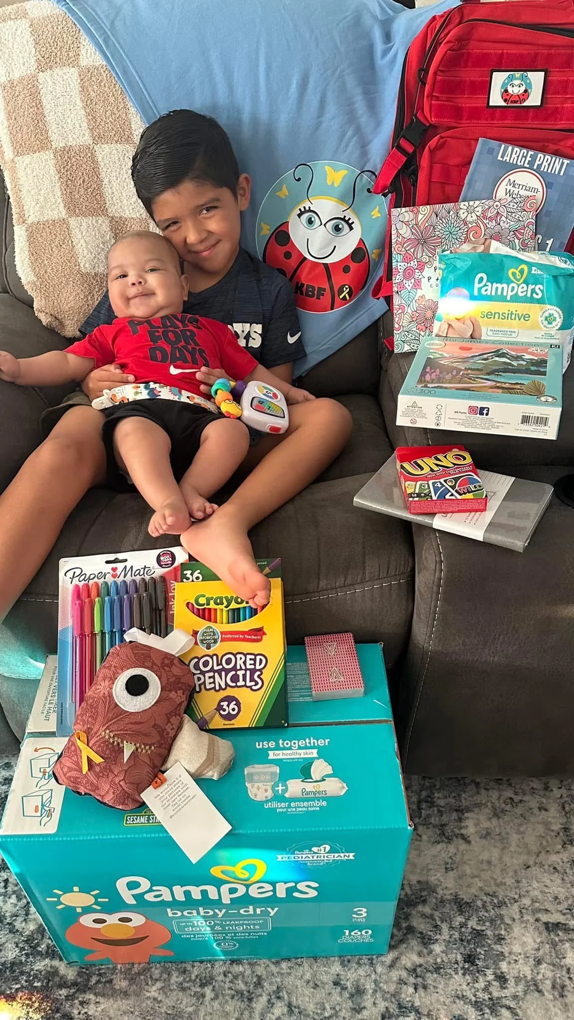 Two children sitting on a couch surrounded by baby supplies, toys, and clothing, including a box of Pampers diapers, coloring supplies, a backpack, and board games.