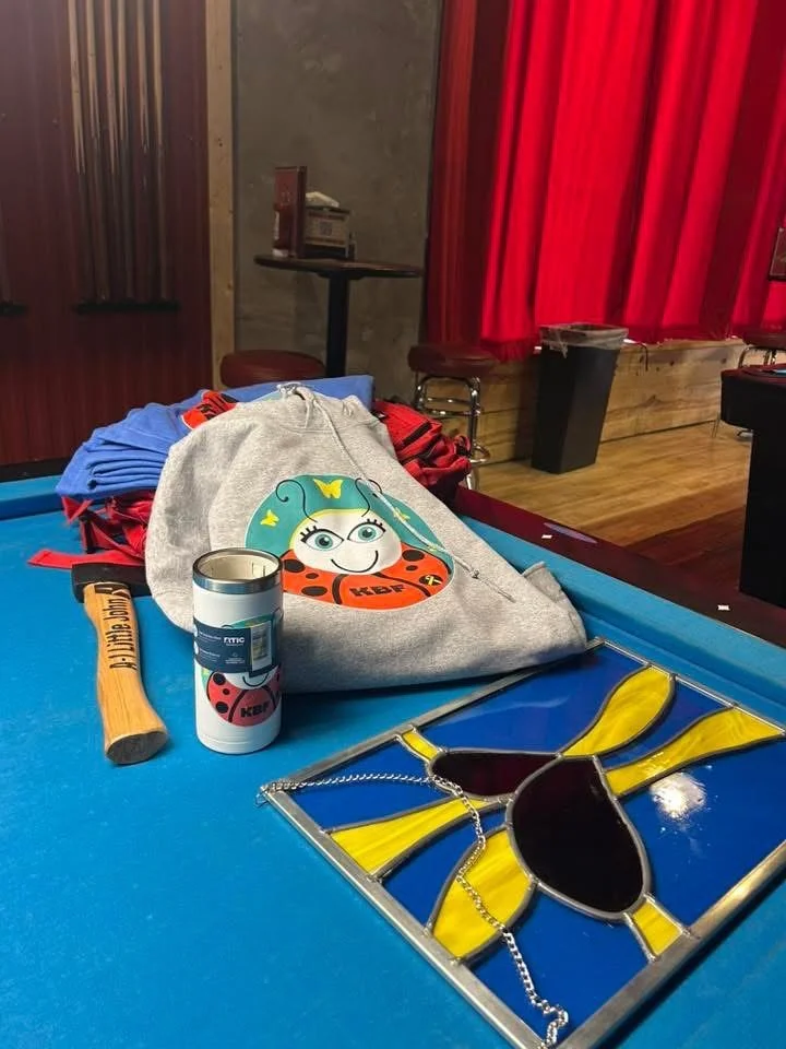 Items on a pool table, including a gray sweatshirt with a ladybug and ladybug-themed graphic, a small metal container, a wooden hammer with "Little Buddy" written on it, a blue object, and a stained glass piece with yellow rays and a honey jar design